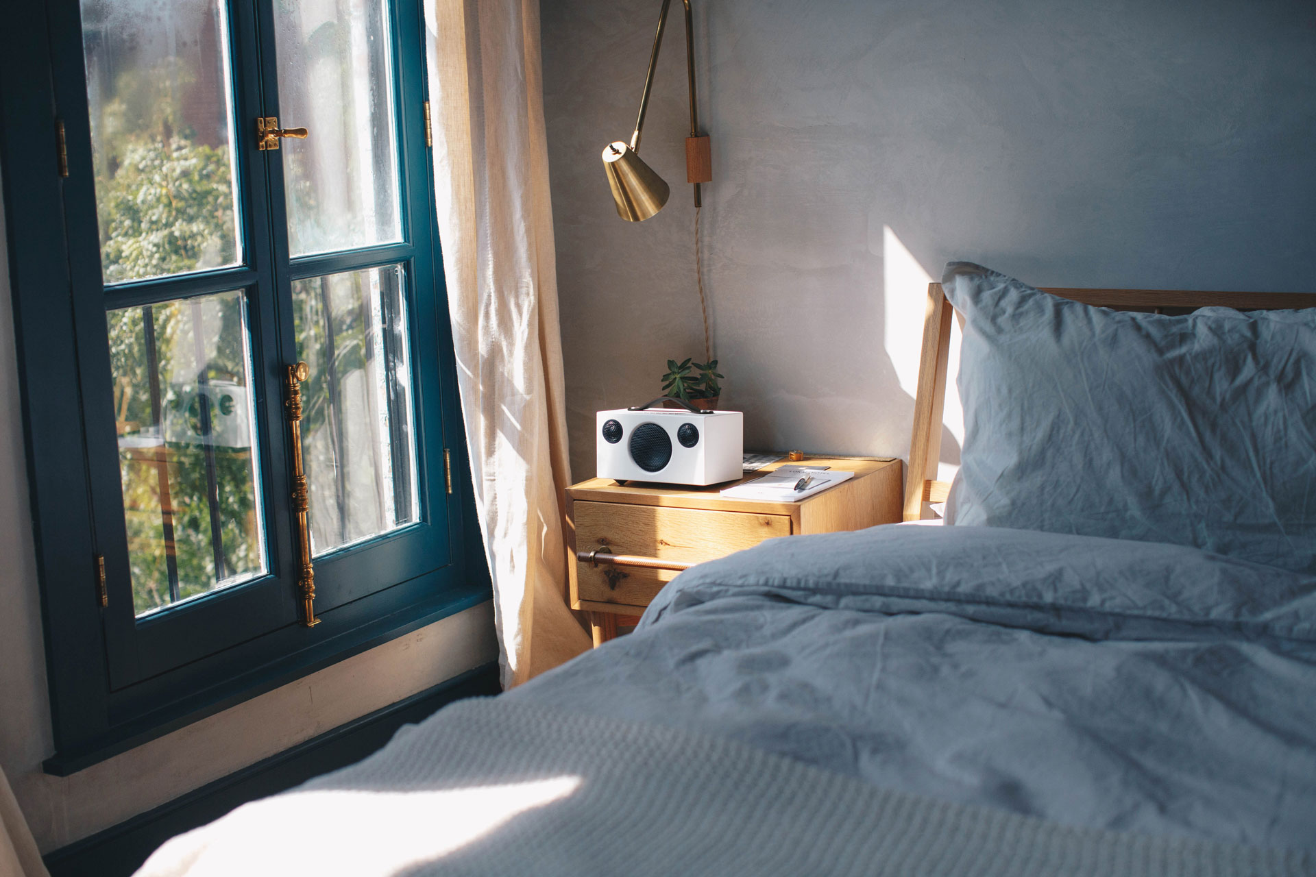 Slaapkamer scène. Bed met lichtgrijs beddengoed. Houten nachtkastje met lamp, plant en een witte speaker. Raam.