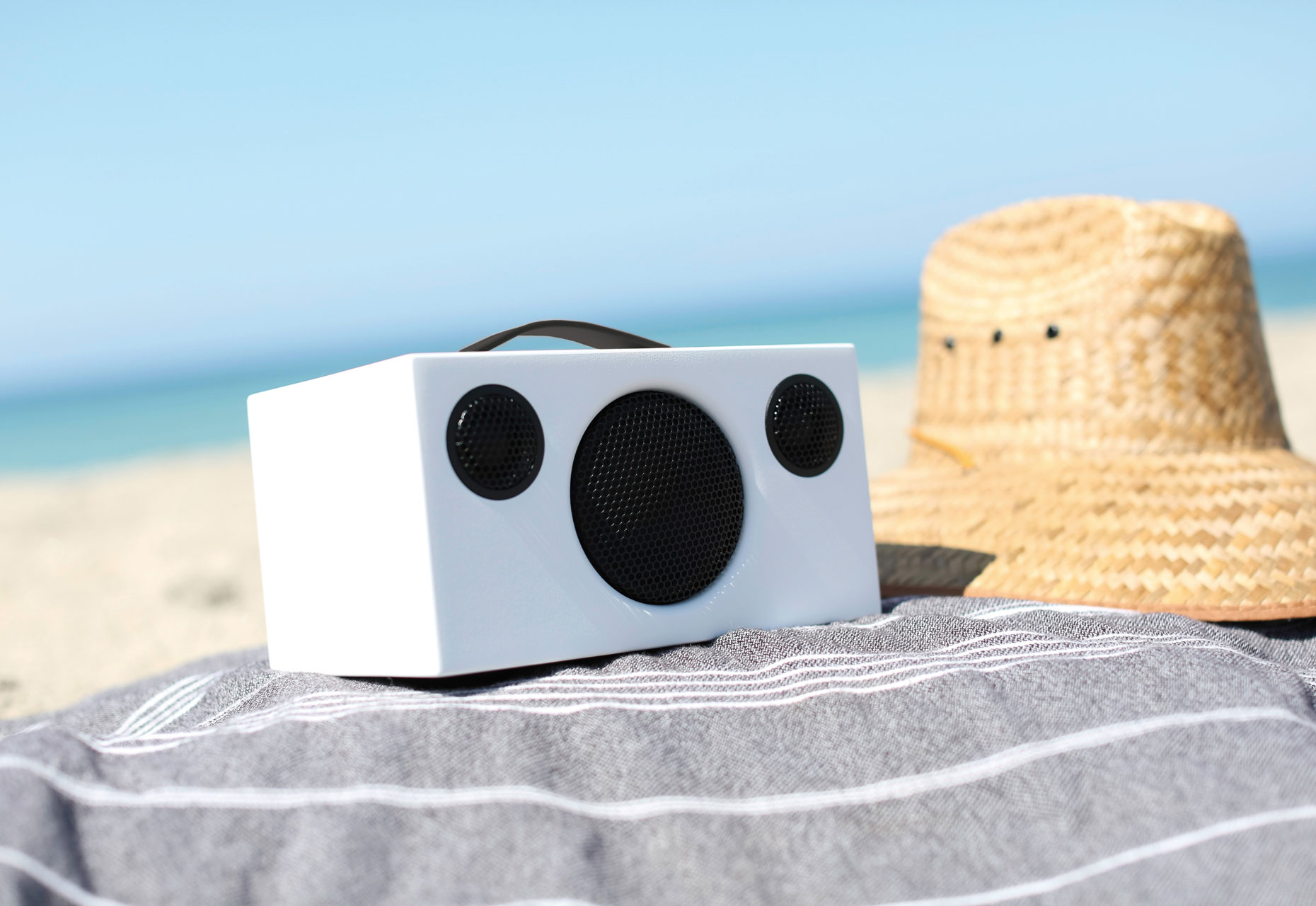 Witte speaker met zwarte speakerroosters op een strandlaken. Strooien hoed rechts.
