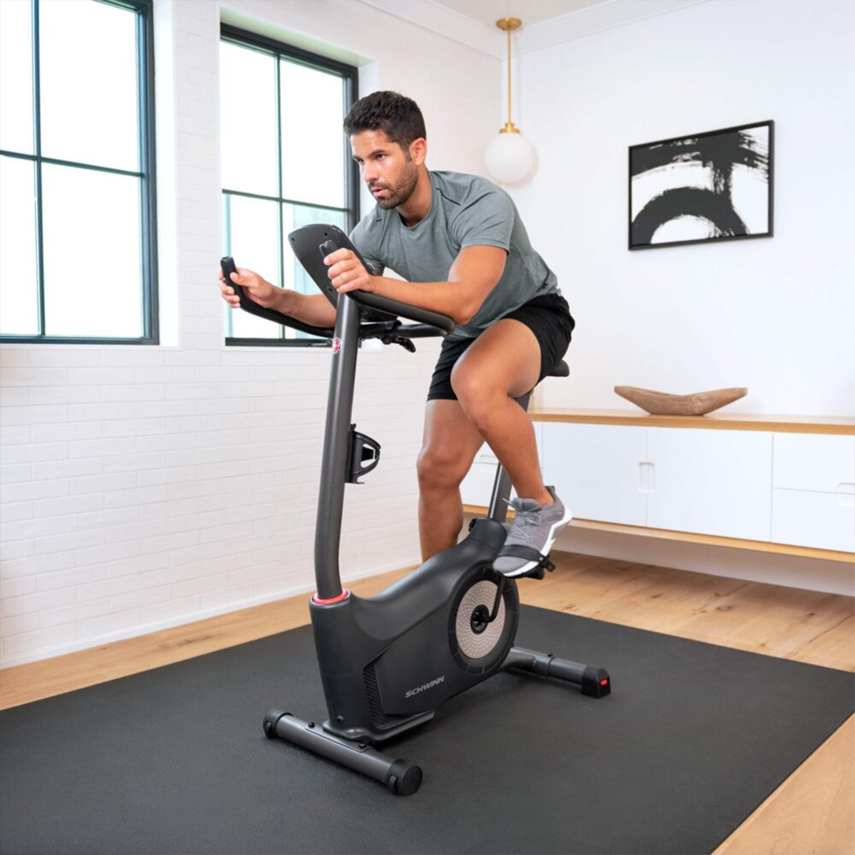 Homme sur un vélo d'appartement, faisant de l'exercice sur un tapis. La pièce a une fenêtre et un tableau.