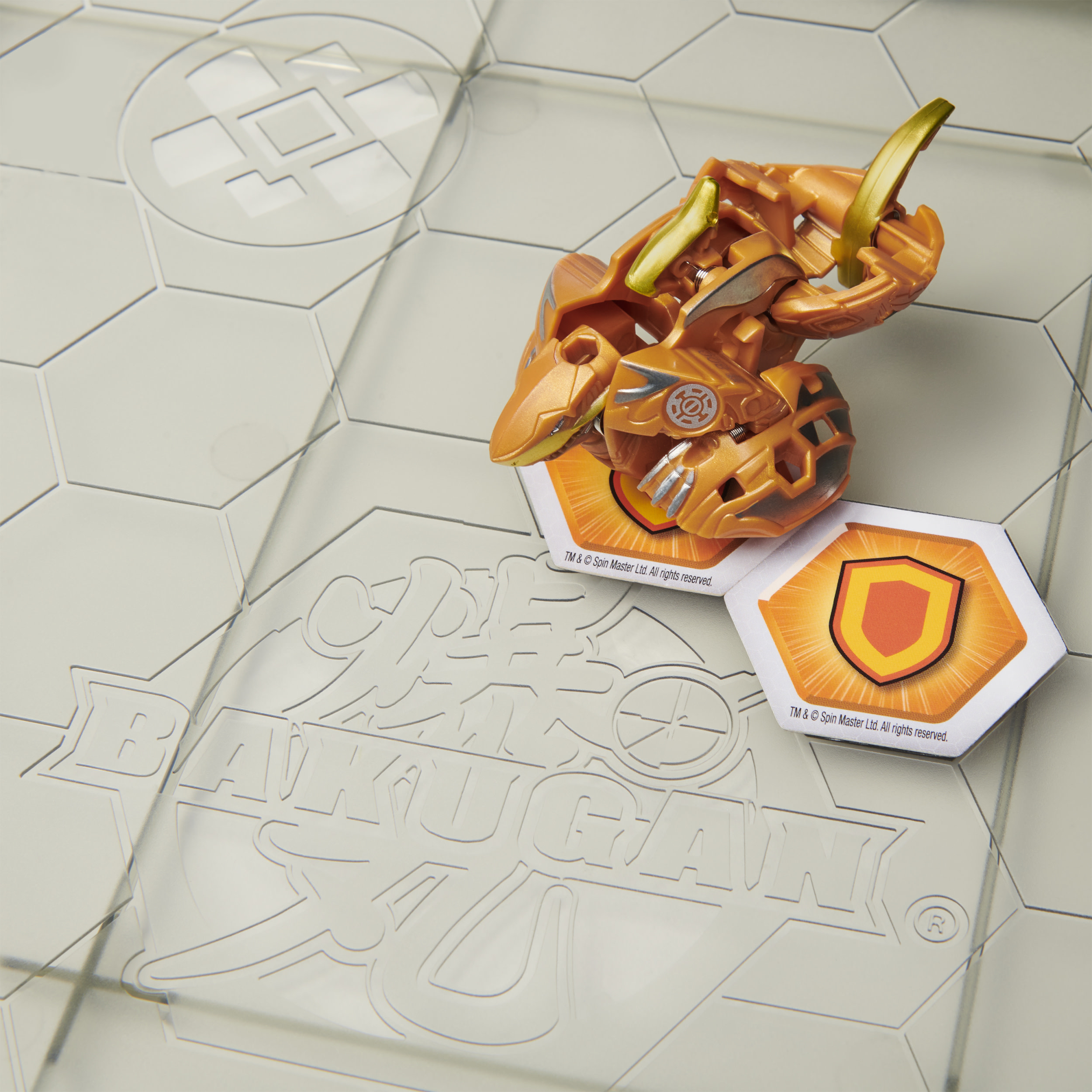 Jouet Bakugan doré et orange sur un tapis de jeu hexagonal. Le Bakugan a deux cartes à proximité.