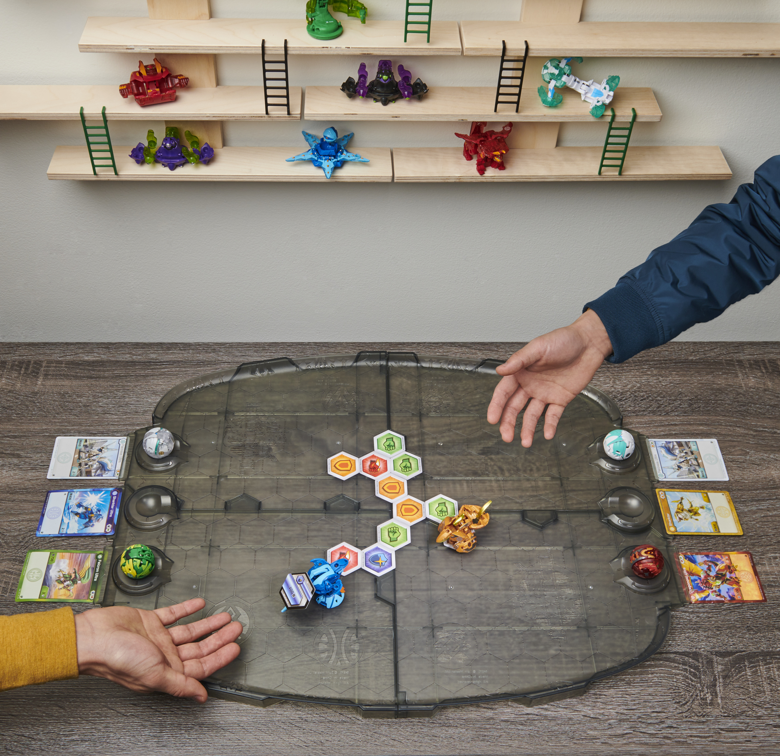Des personnes jouant avec des figurines sur un plateau de jeu. Figurines sur des étagères en arrière-plan. Mains qui se tendent vers le plateau.