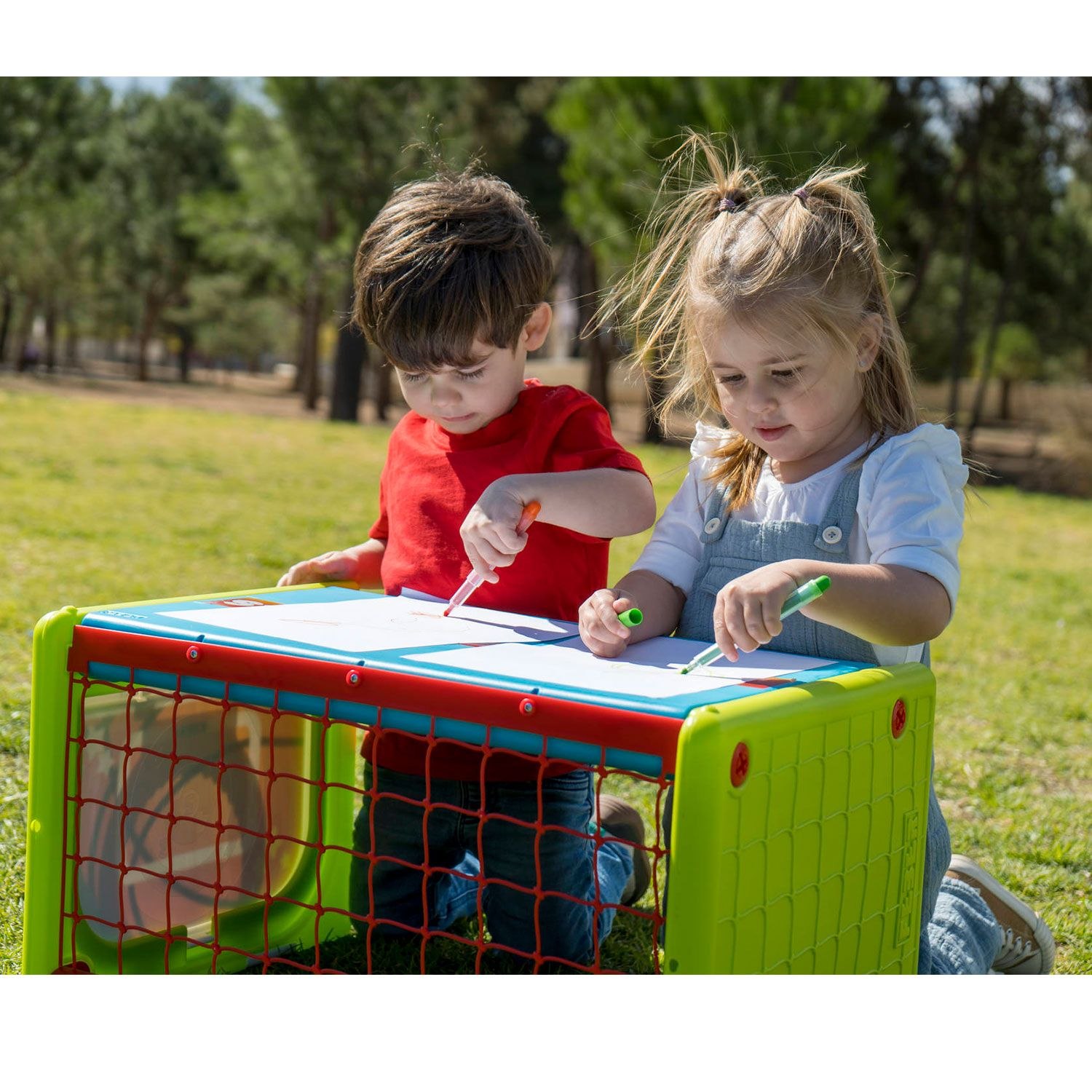 Due bambini disegnano a un tavolo verde con una rete rossa su un campo erboso con alberi.