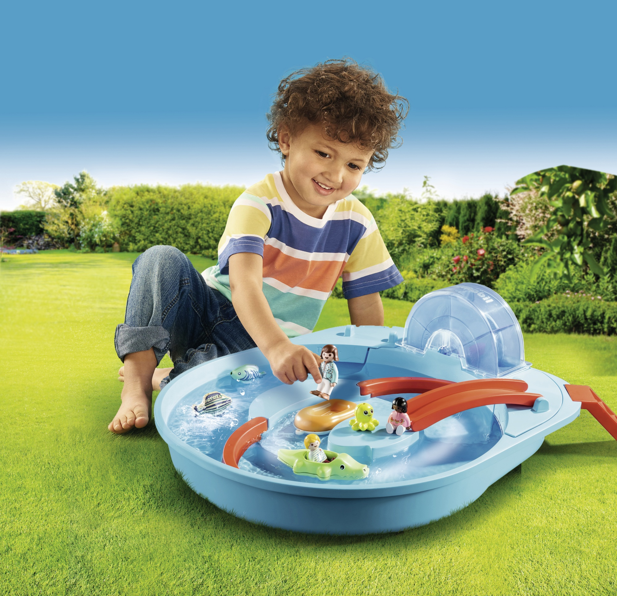 Un bambino che gioca con un set di parco acquatico giocattolo, con personaggi e accessori. La scena è ambientata all'aperto sull'erba.