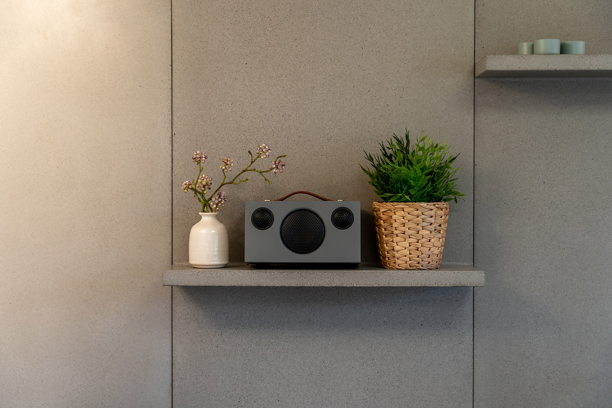 Een grijze luidspreker op een plank met een vaas bloemen en een plant in een pot.