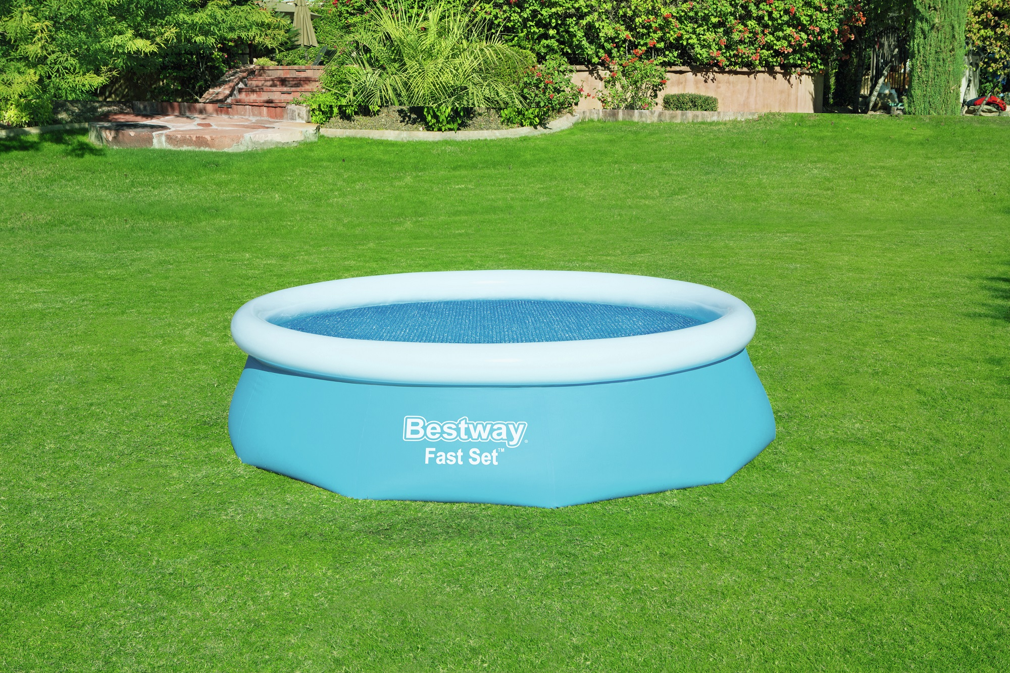 Piscina inflable azul sobre césped verde, con borde blanco y fondo de árboles y una casa.