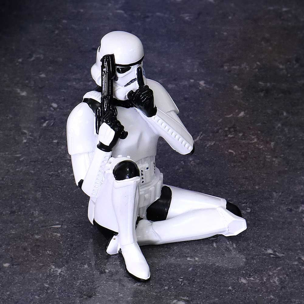 Stormtrooper figuur, zittend op grijs oppervlak, houdt een blaster vast, wijsvinger op lippen, witte armor, zwarte handschoenen.