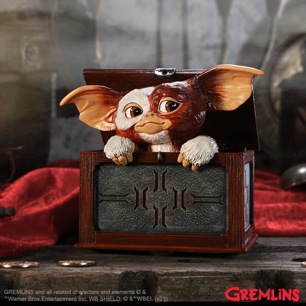 Een Gremlin-figuur kijkt uit een bruine houten kist met een rode achtergrond. De figuur heeft grote oren en witte vacht.