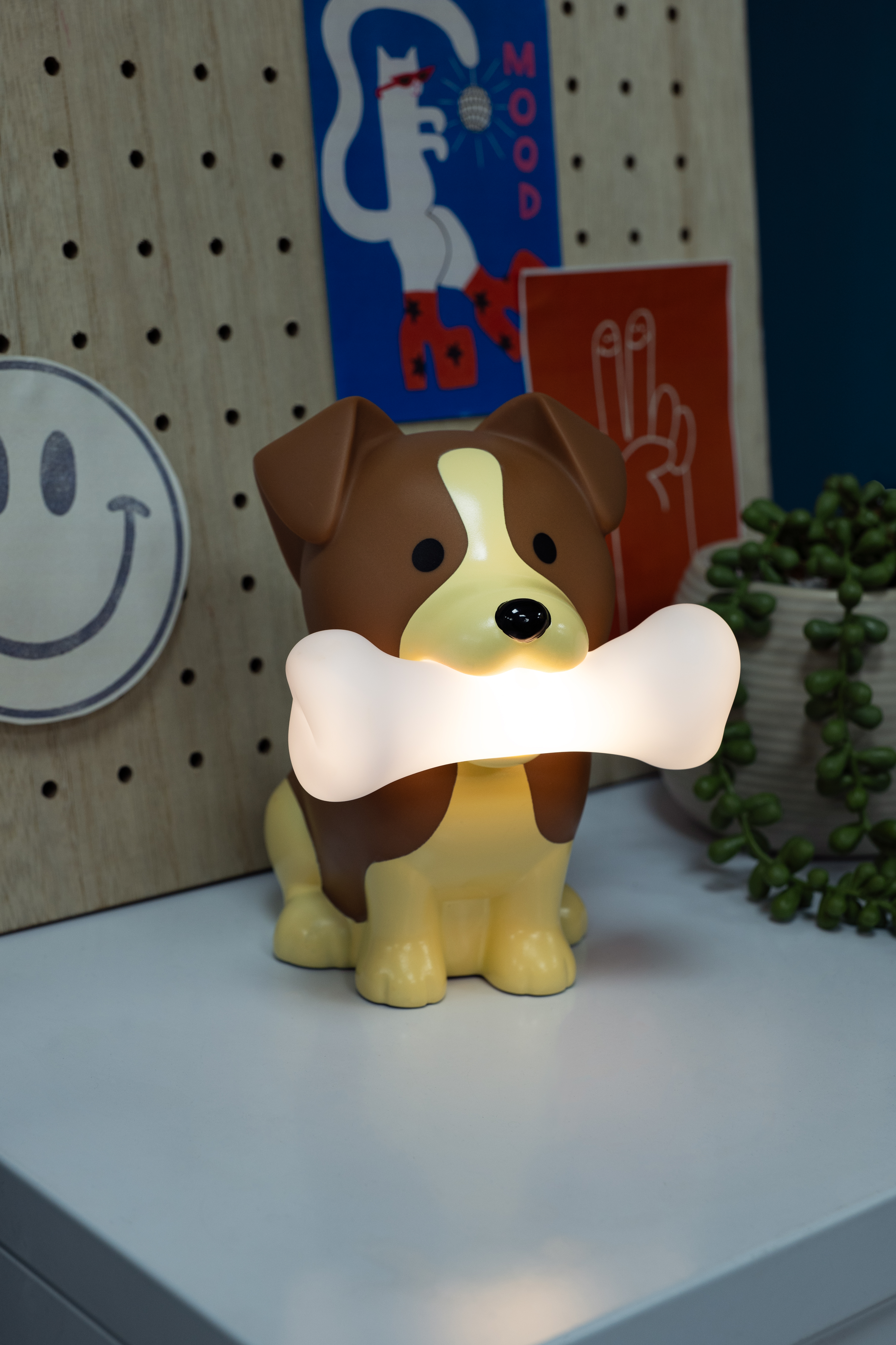 A dog-shaped lamp holding a bone. Background has drawings and a plant pot.