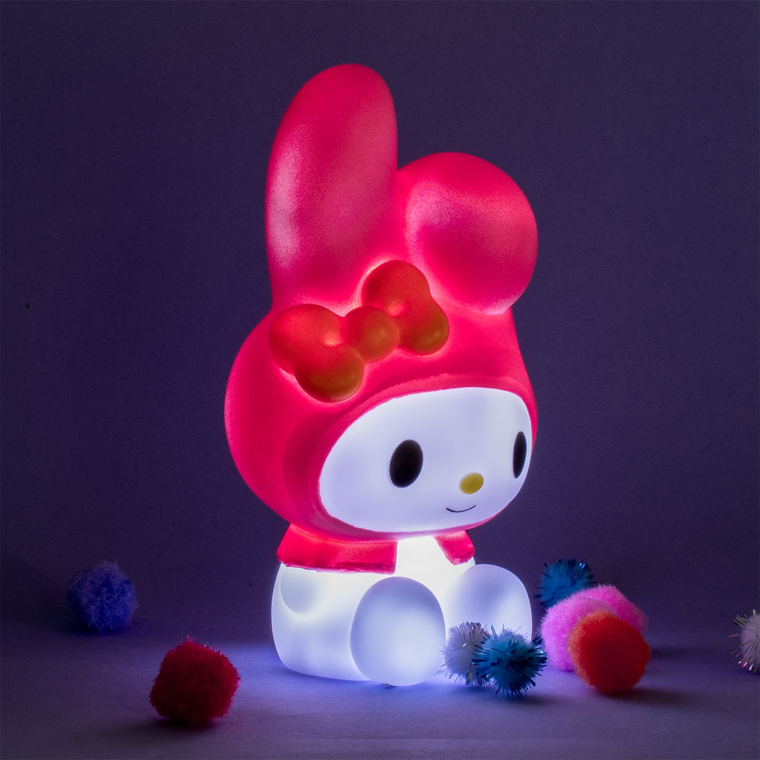 Een roze My Melody lamp. Hij is verlicht en heeft een rode strik. Er liggen pompons in de buurt.
