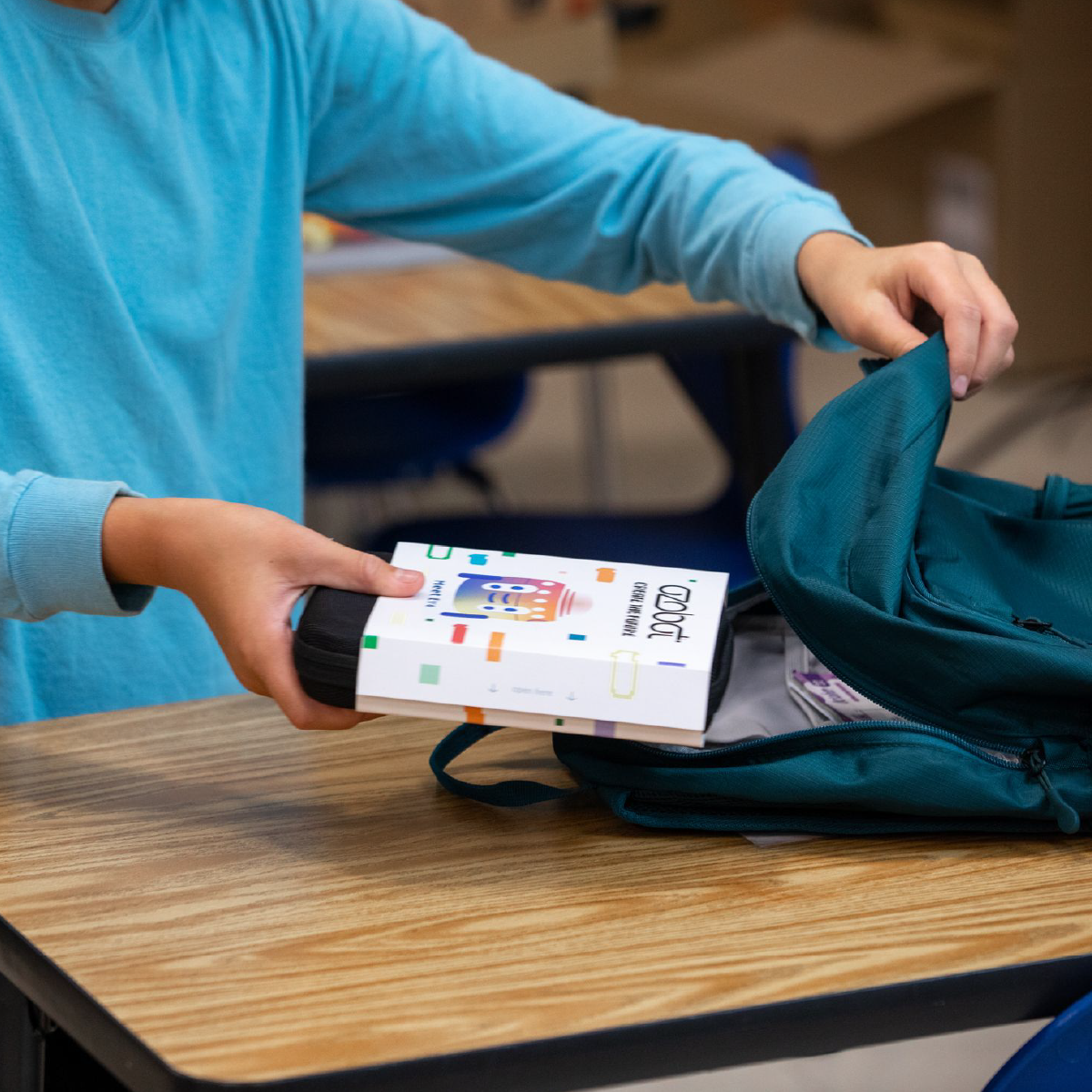 A person putting a colorful box into a teal backpack on a desk.