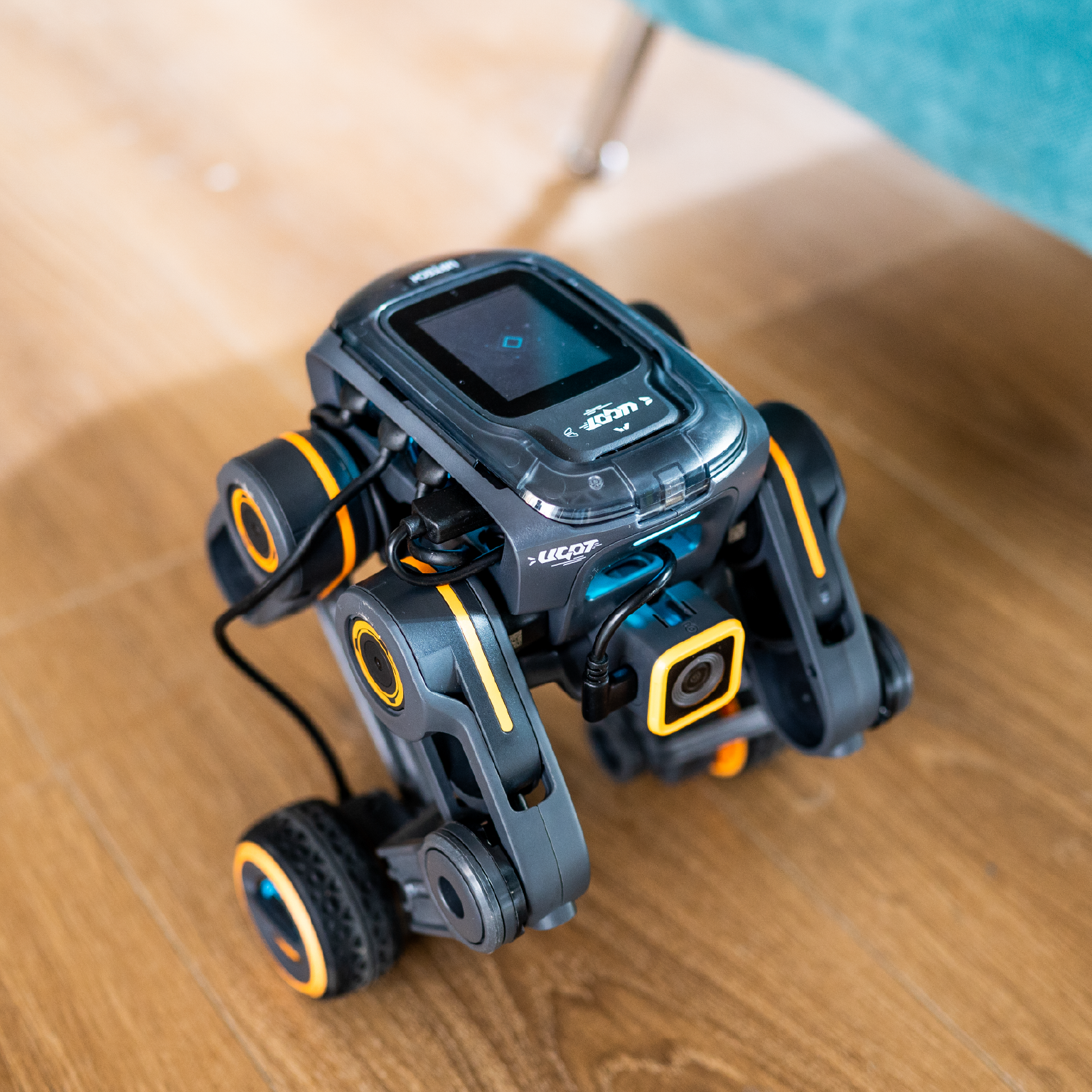 A robot with orange and gray colors. The robot is on a wooden floor.