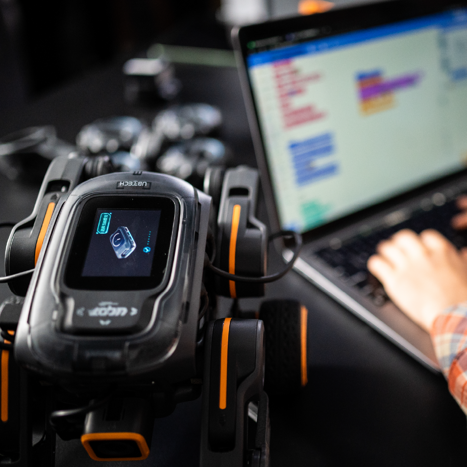 A robot with a screen displaying an image. A person is using a laptop with a programming code.