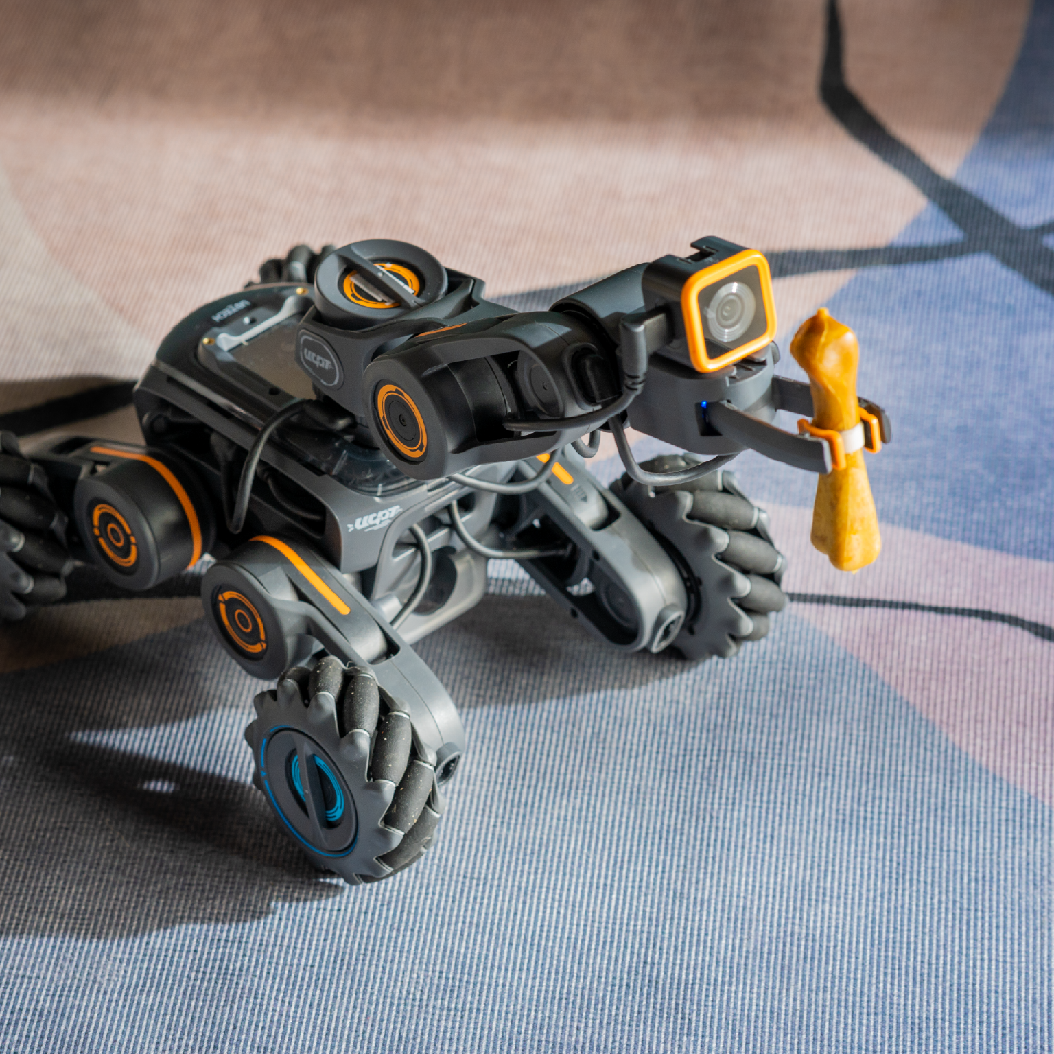Un vehículo robot gris con acentos naranjas, una cámara y una garra que sostiene un objeto amarillo.