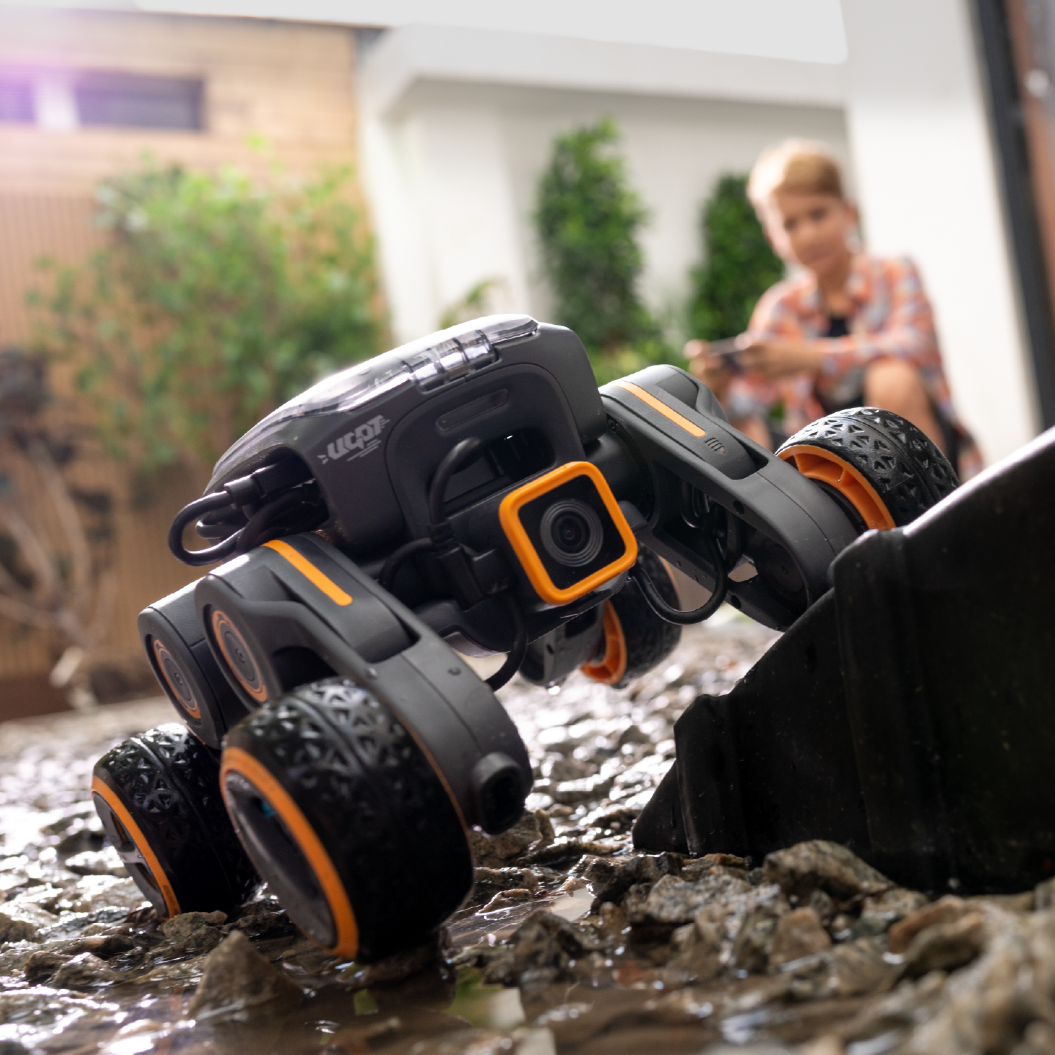 Coche teledirigido negro y naranja sobre rocas. Niño con teléfono al fondo.