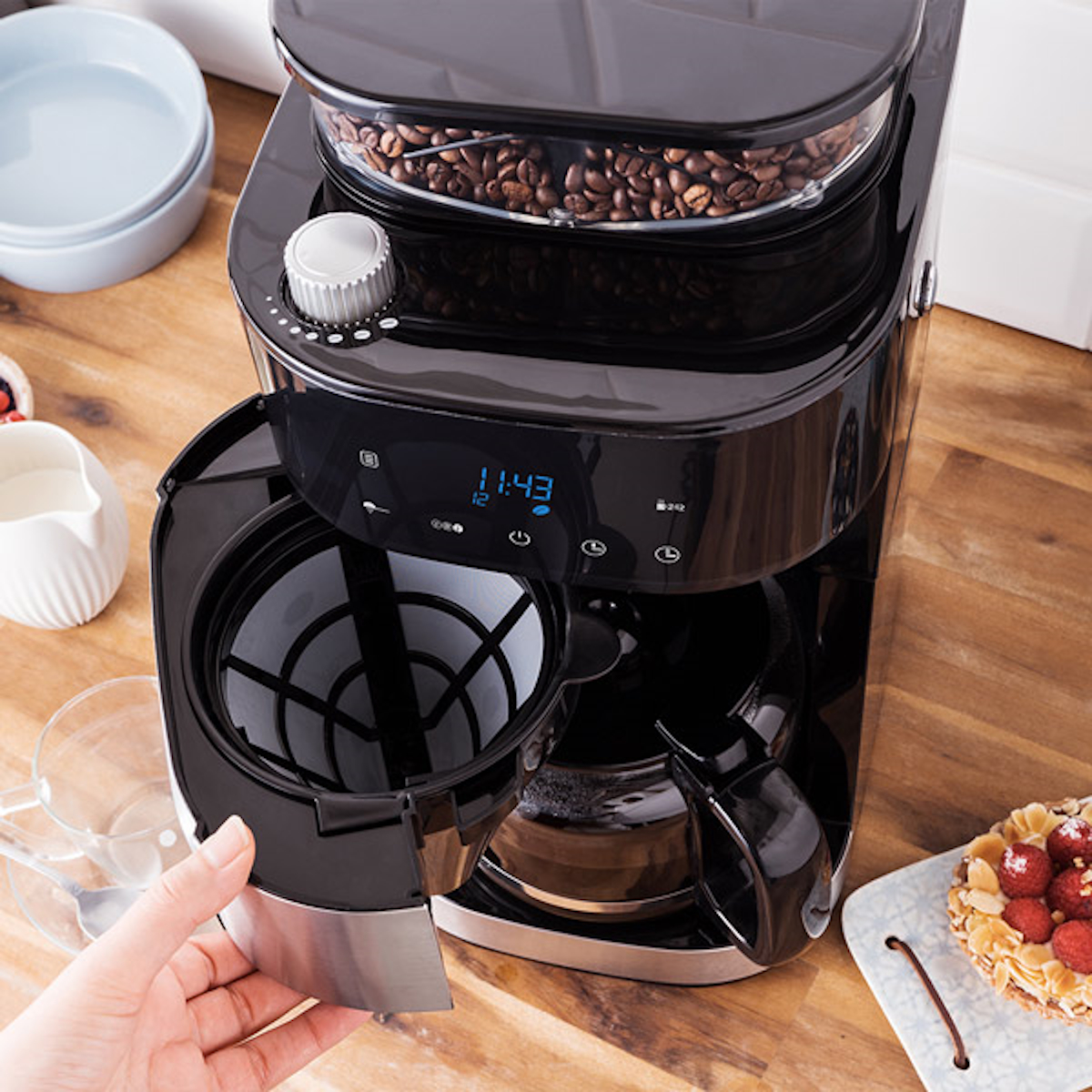Czarny ekspres do kawy z ziarnami kawy w środku, otwartym koszem filtra i ręką sięgającą po niego.