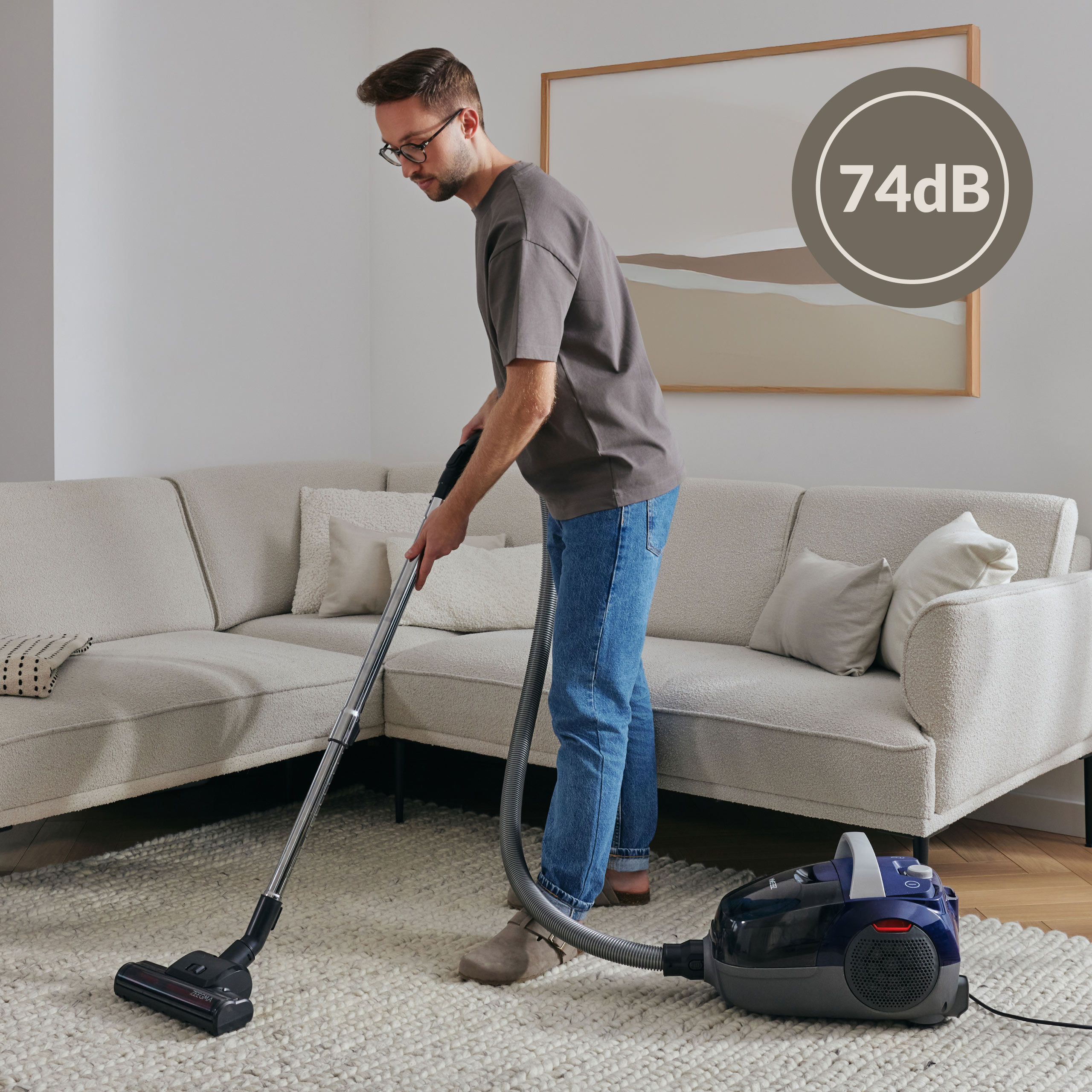 Mężczyzna odkurza dywan w salonie, 74dB, sofa i obraz.