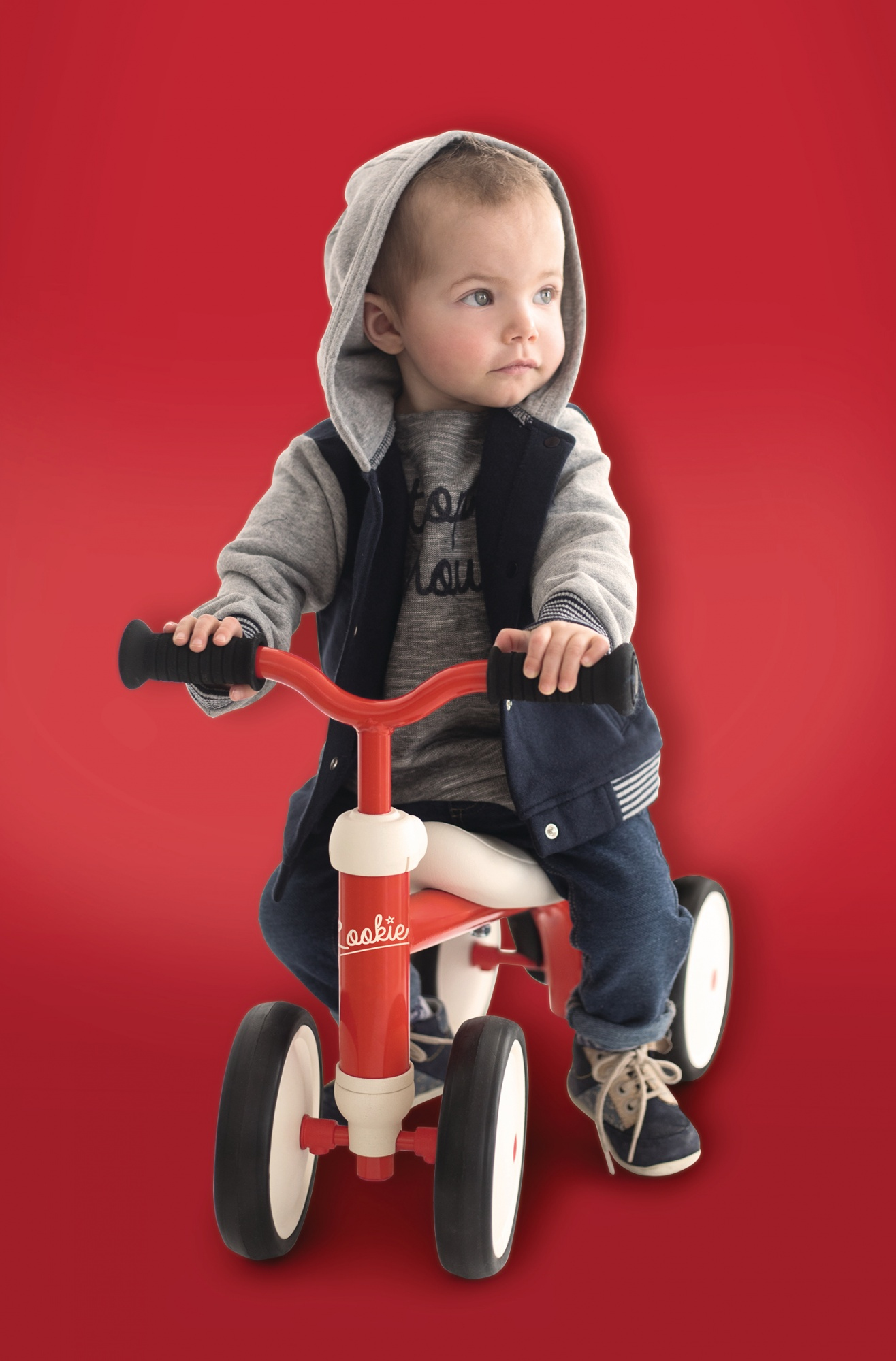Dziecko na czerwonym rowerku, w kurtce i bluzie z kapturem, na czerwonym tle.