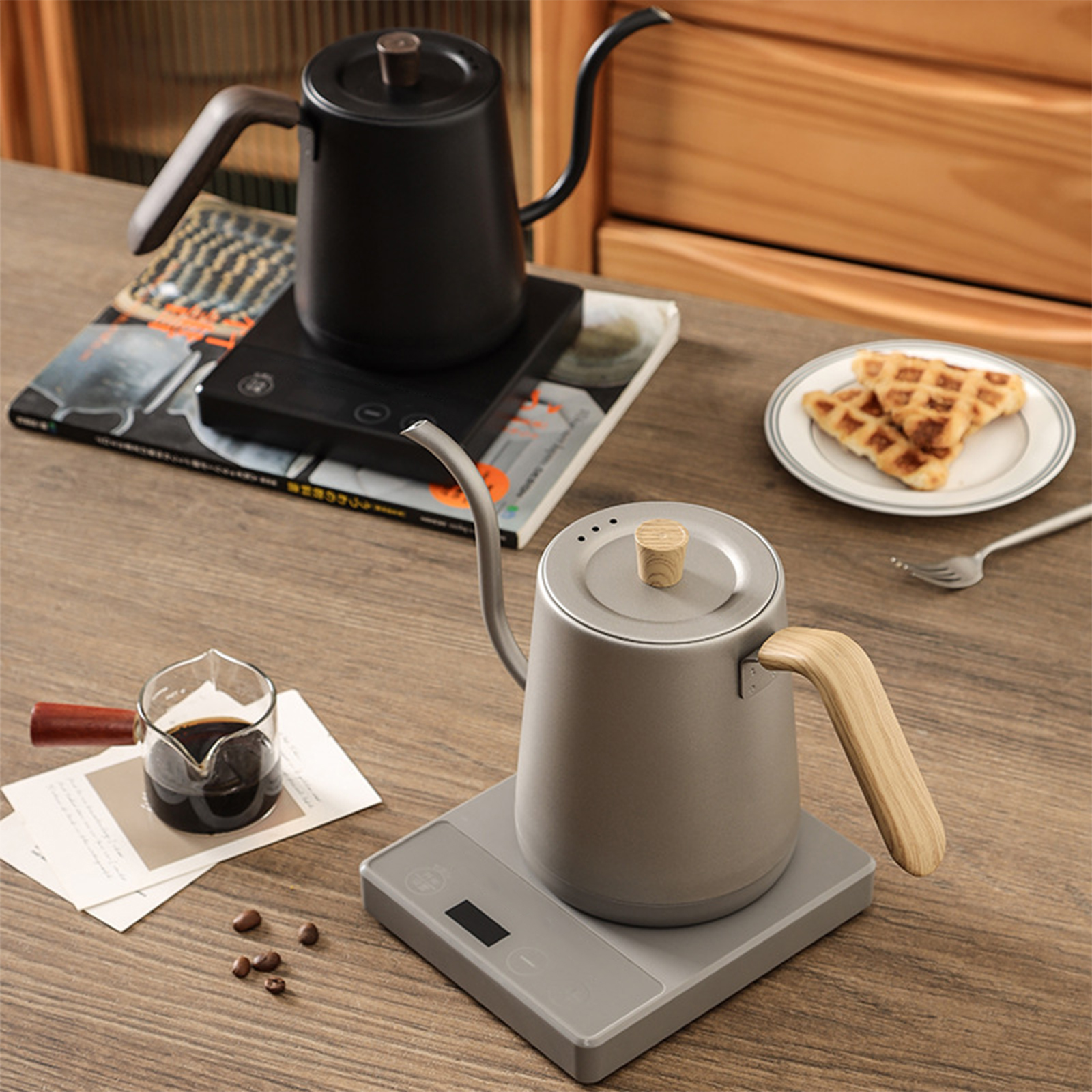 Zwei Wasserkocher auf elektrischen Sockeln, einer schwarz und einer grau, mit einer Tasse Kaffee und Waffeln.