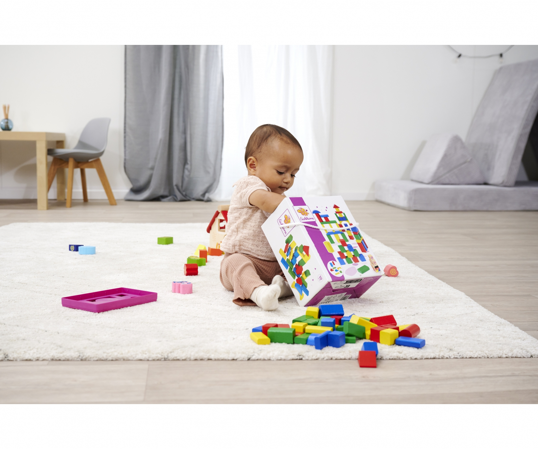 Ein Baby spielt mit bunten Bausteinen aus einer Kiste auf einem Teppich. Spielzeug liegt herum.