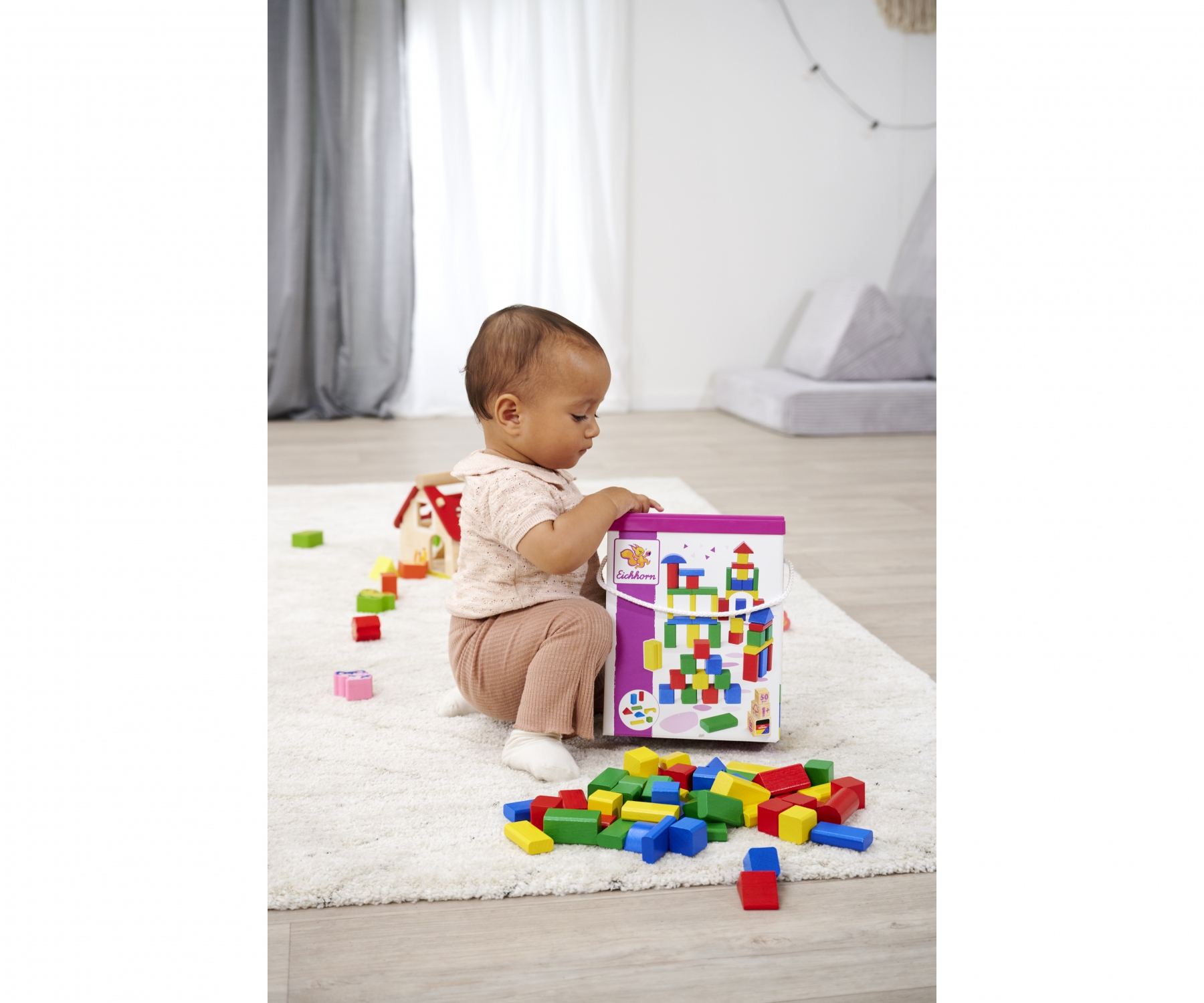 Ein Baby sitzt auf einem Teppich, umgeben von bunten Spielzeugblöcken und einer Kiste mit Blöcken.