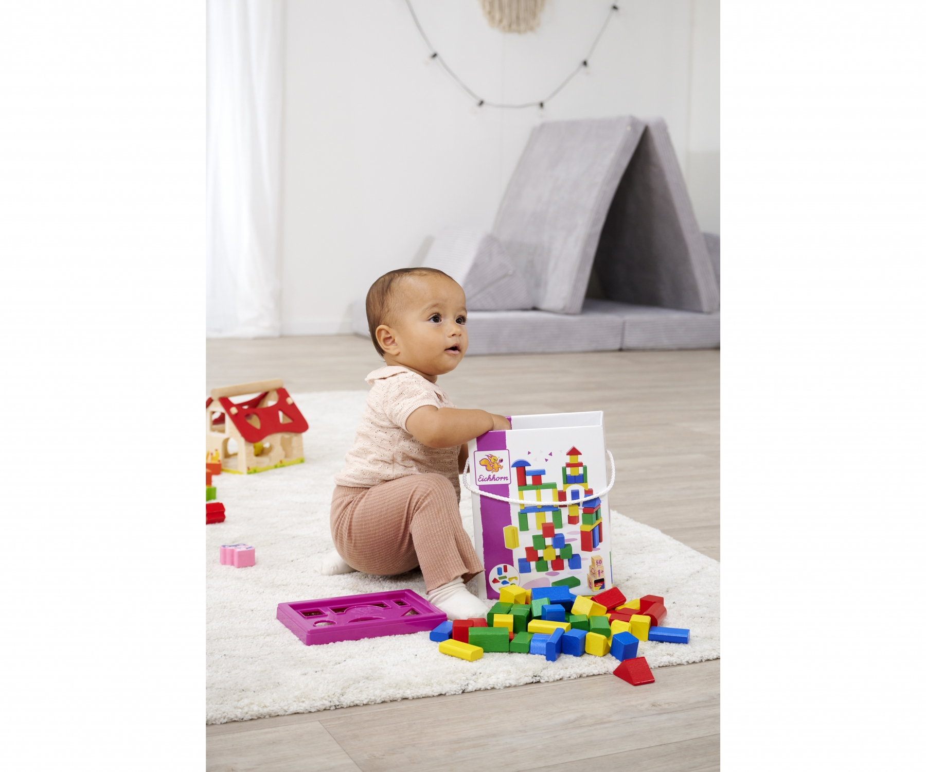 Ein Baby sitzt mit Blöcken und einem Spielzeughaus. Spielzeug und eine Matte liegen auf dem Boden.