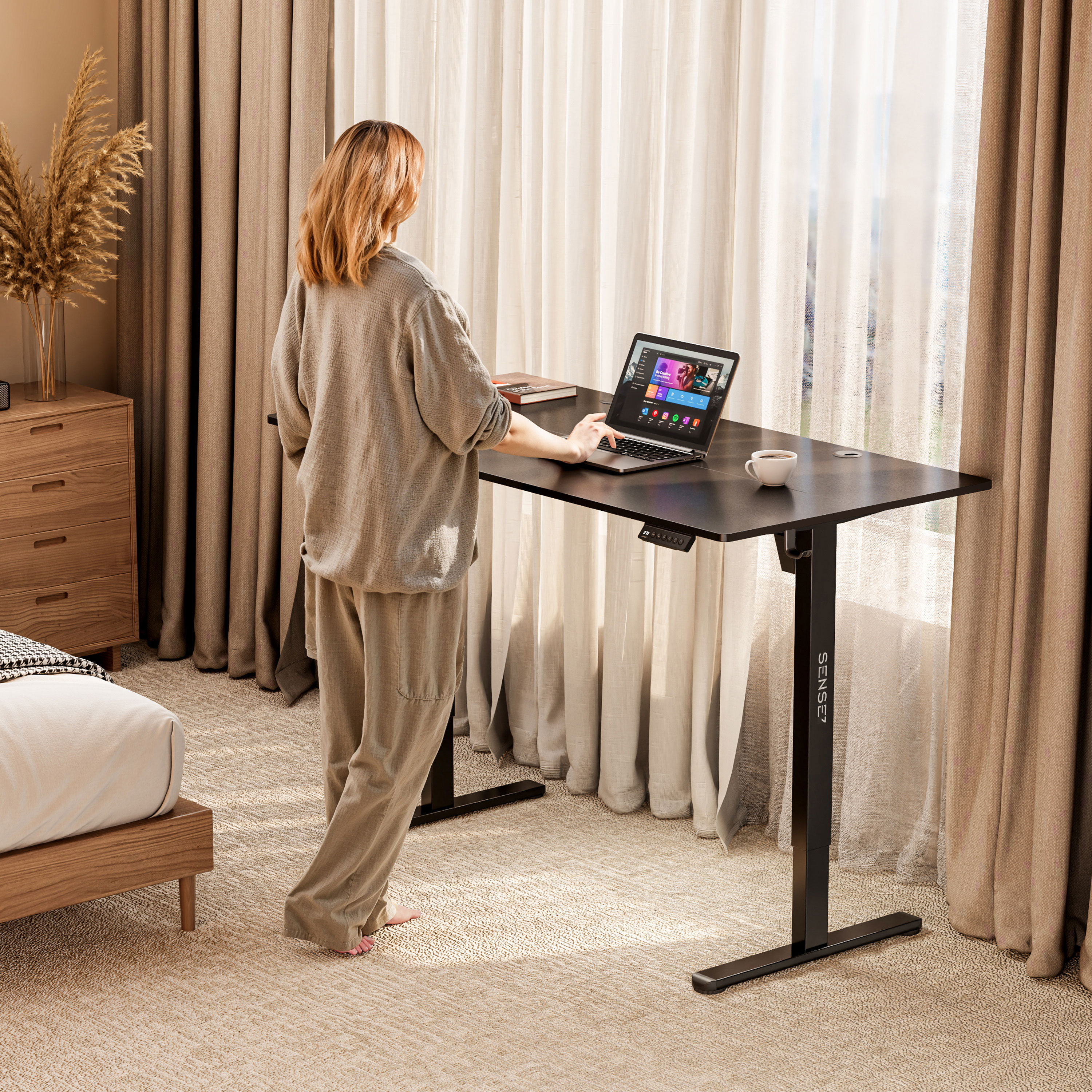 Donna in piedi a una scrivania nera con un laptop. Un letto e una finestra sono sullo sfondo.