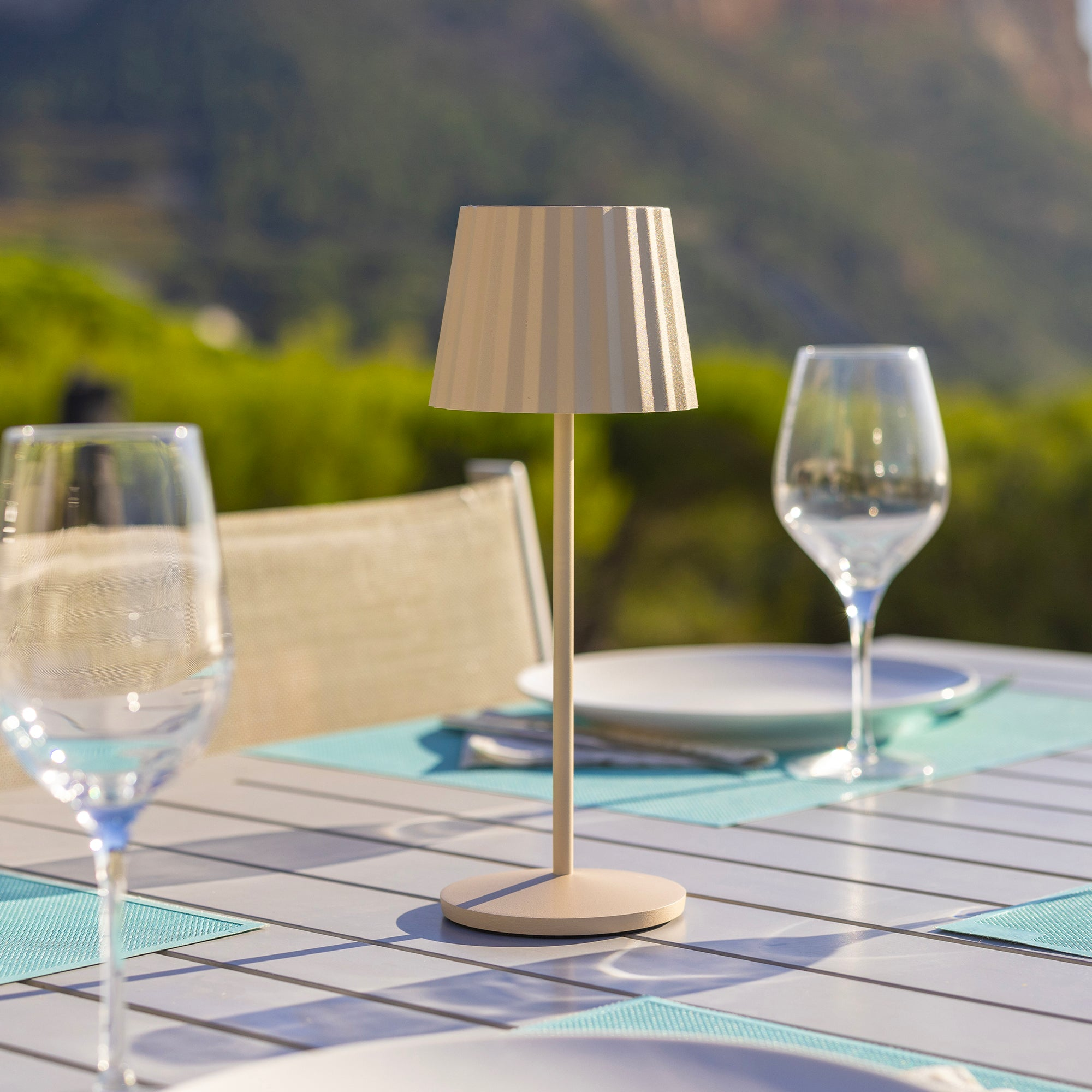 Eine Tischlampe mit cremefarbenem Schirm auf einem Tisch mit Weingläsern und Blick auf eine grüne Landschaft.