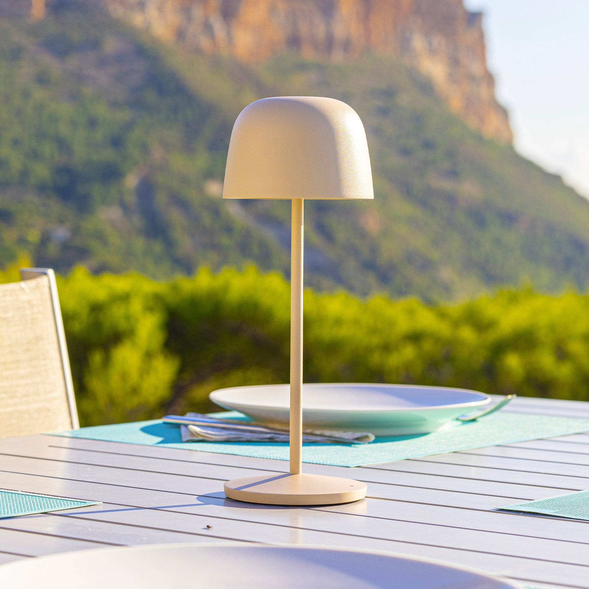 Eine Tischlampe auf einem Tisch im Freien. Die Lampe ist beige, im Hintergrund sind Berge und grüne Bäume zu sehen.
