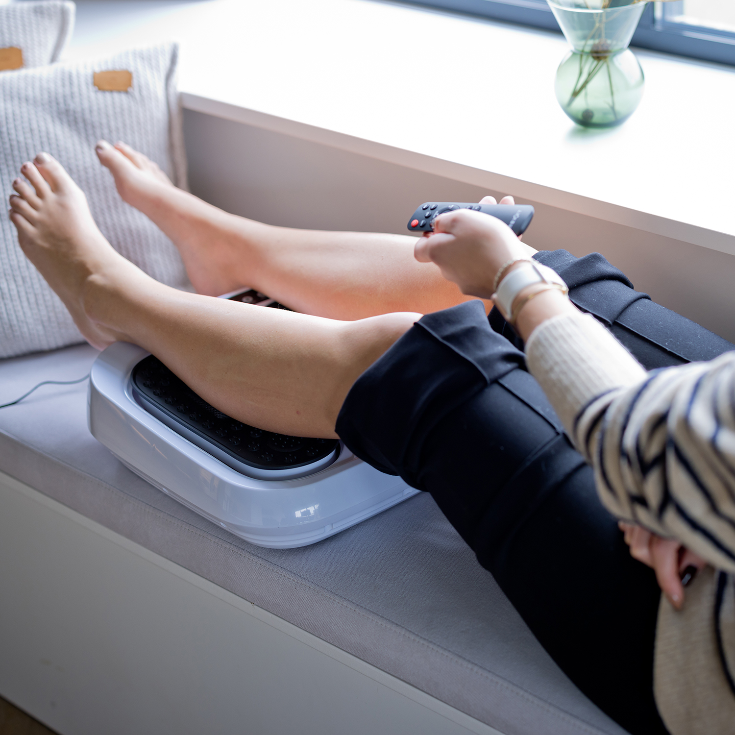 A person is using a foot massager while resting on a couch. They are holding a remote.