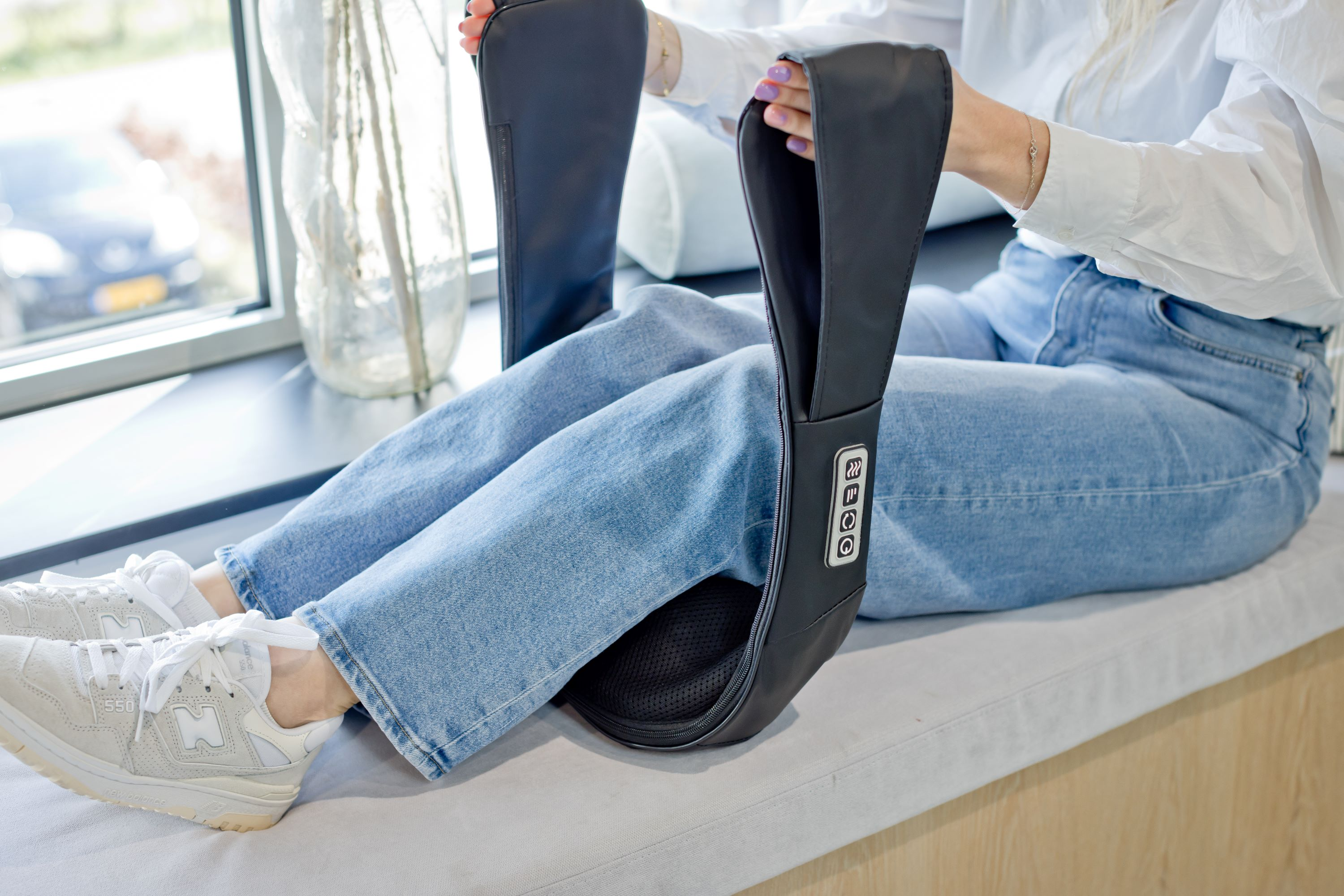 Woman in jeans sitting with a black massage device on her shoulders.