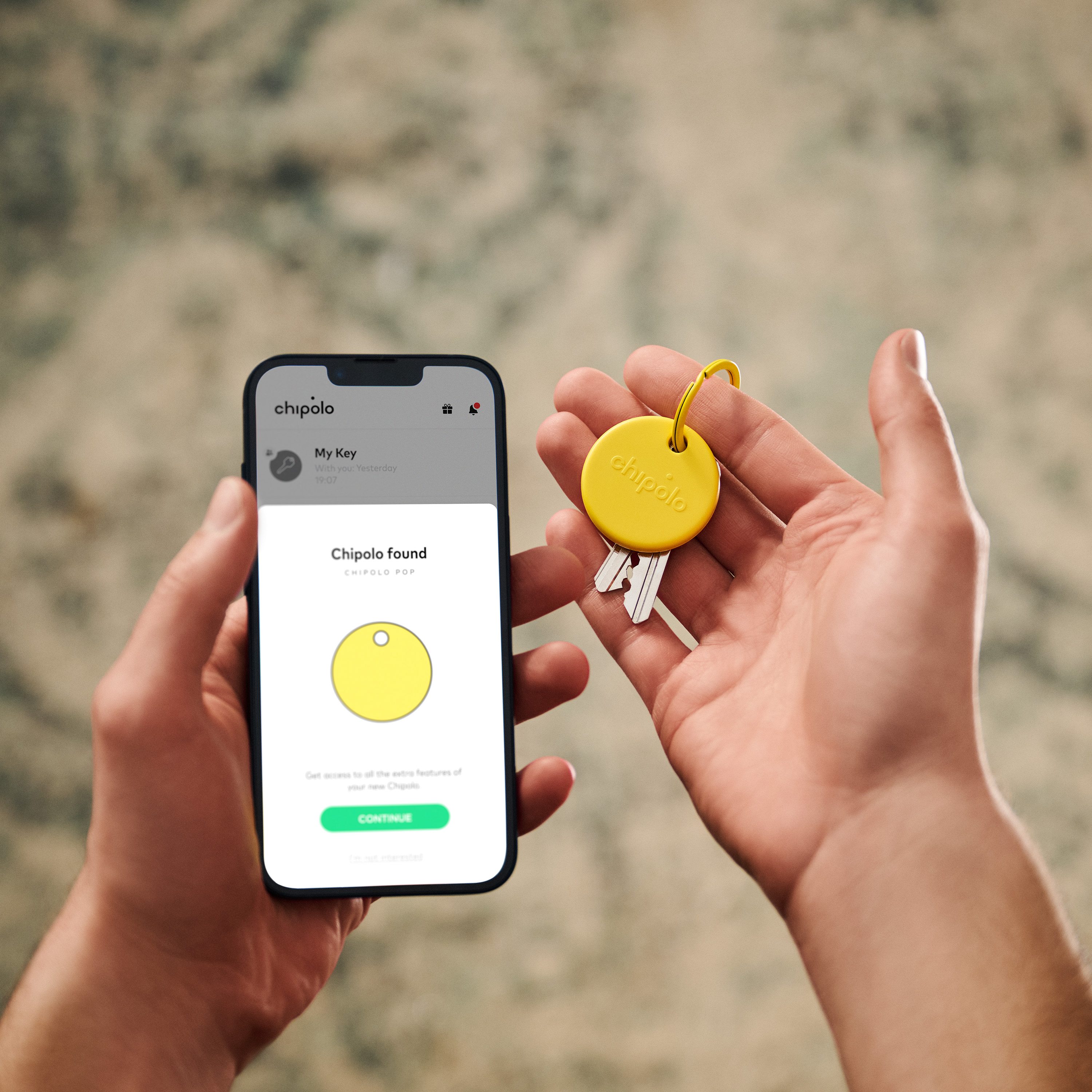 Person holding phone with Chipolo app. Yellow Chipolo tag with keys. Blurred background.