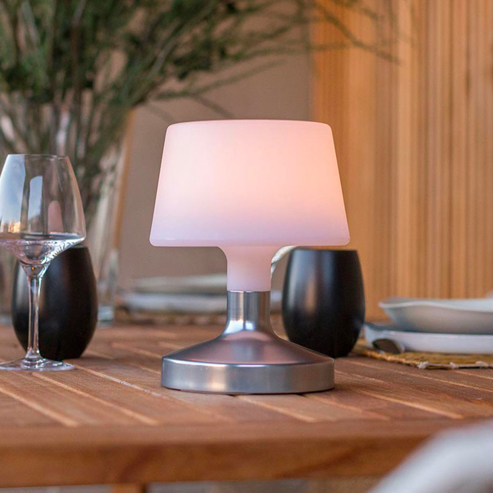 Eine beleuchtete Lampe mit weißem Schirm und silbernem Fuß steht auf einem Holztisch.