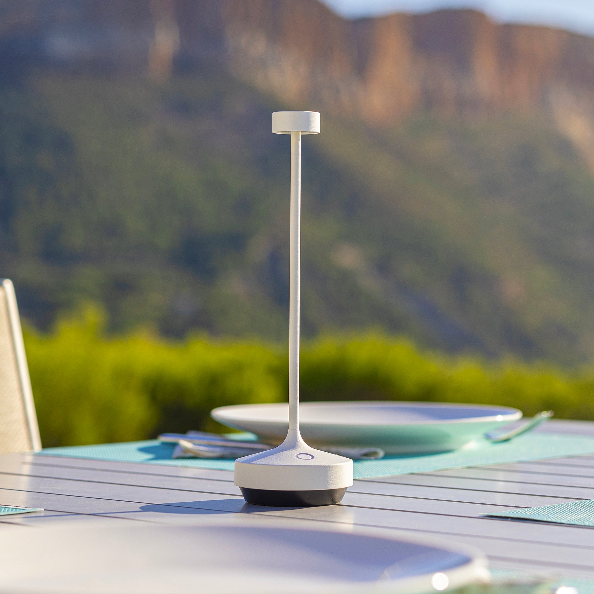 Eine weiß-schwarze Lampe steht auf einem Tisch. Grüne Bäume und ein Berg im Hintergrund.