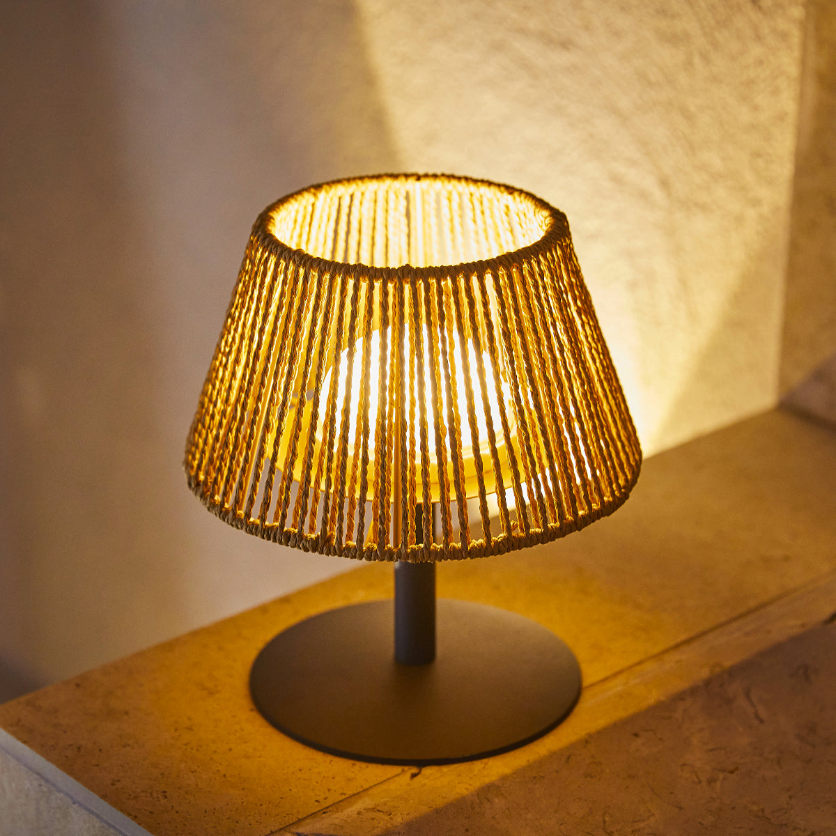 Kleine Korblampe mit schwarzem Sockel, die ein warmes Licht auf einer Holzoberfläche erzeugt.