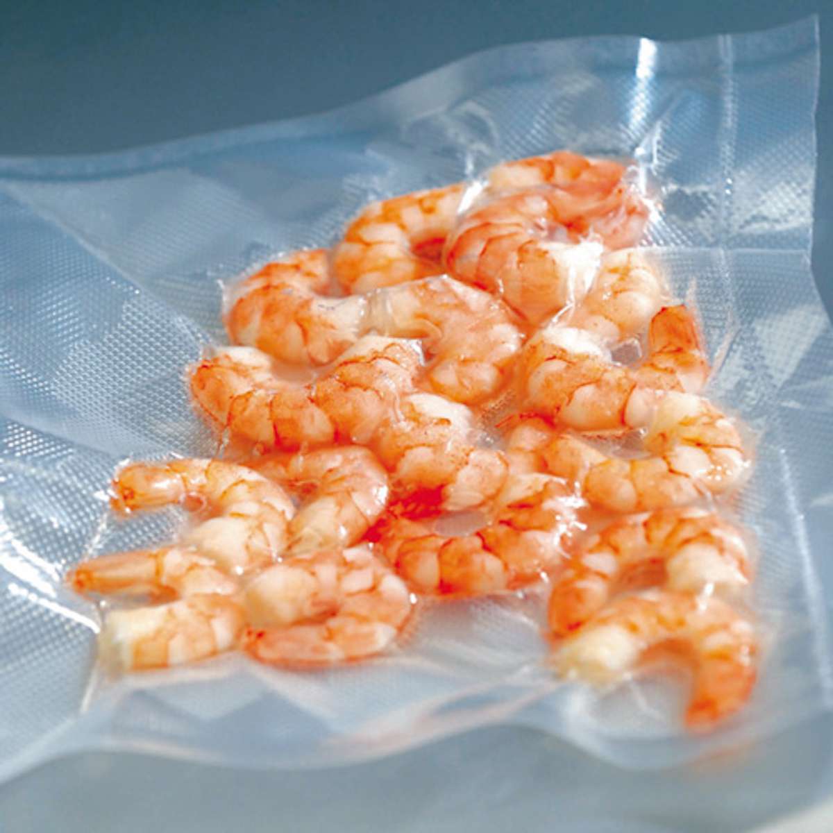 Krewetki zapieczętowane w przezroczystej plastikowej torbie. Są pomarańczowe i różowe. Torba leży na białej powierzchni.