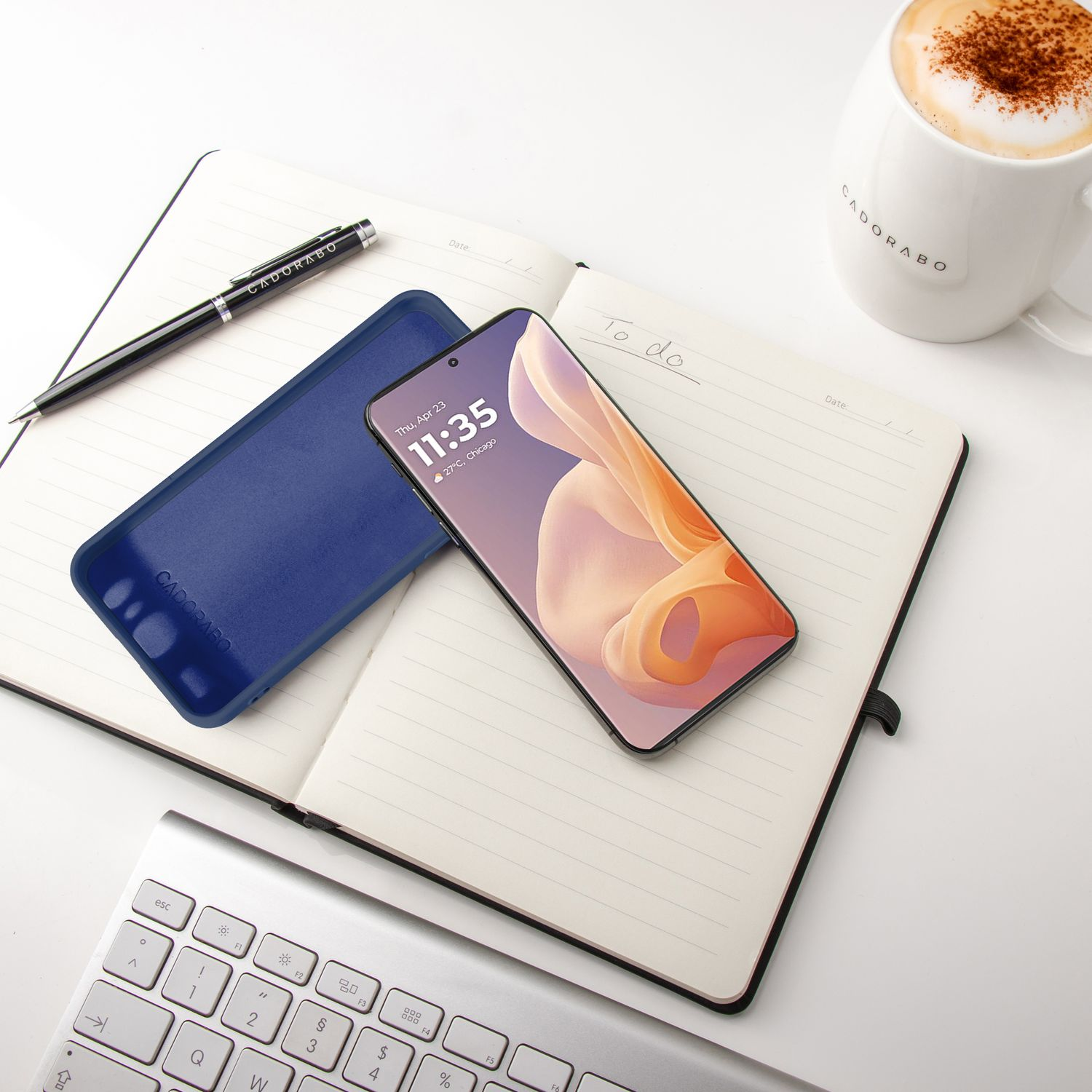 Smartphone auf Notizbuch, Stift und Becher auf weißem Schreibtisch. Hülle neben dem Telefon.