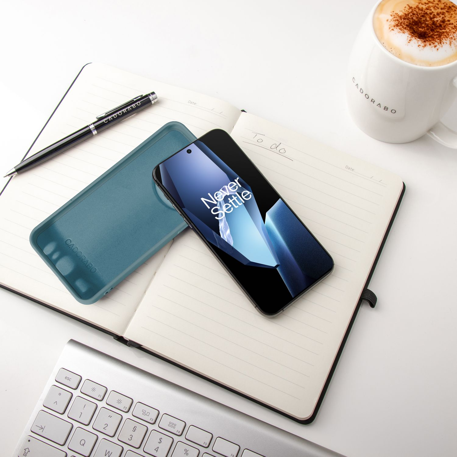 Telefon mit Hülle und Stift auf einem Notizbuch. Eine Tasse Latte und Tastatur sind ebenfalls im Bild.