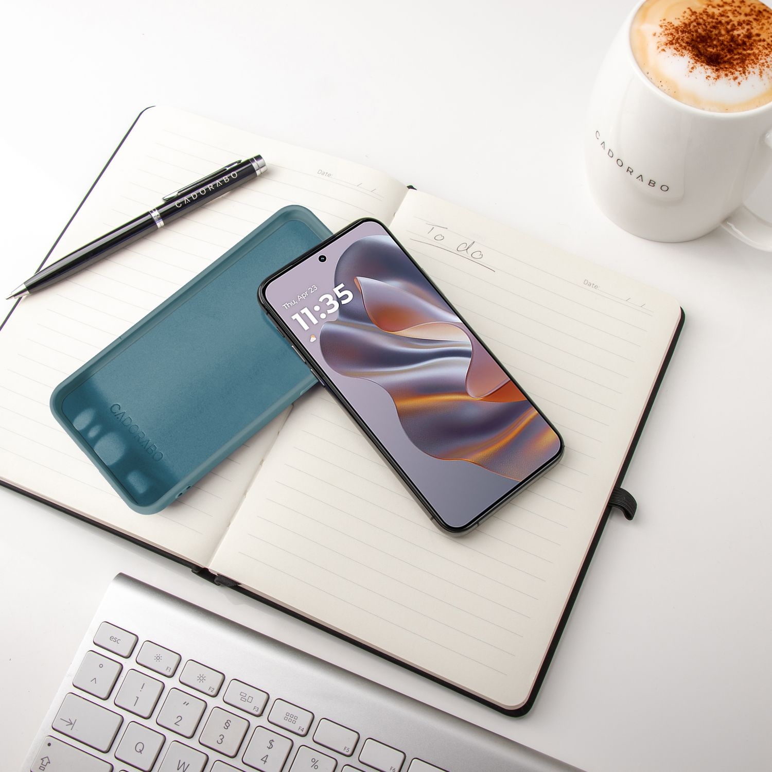 Ein Telefon, eine Hülle, ein Stift und eine Tasse Kaffee liegen auf einem Notizbuch auf einem weißen Tisch.
