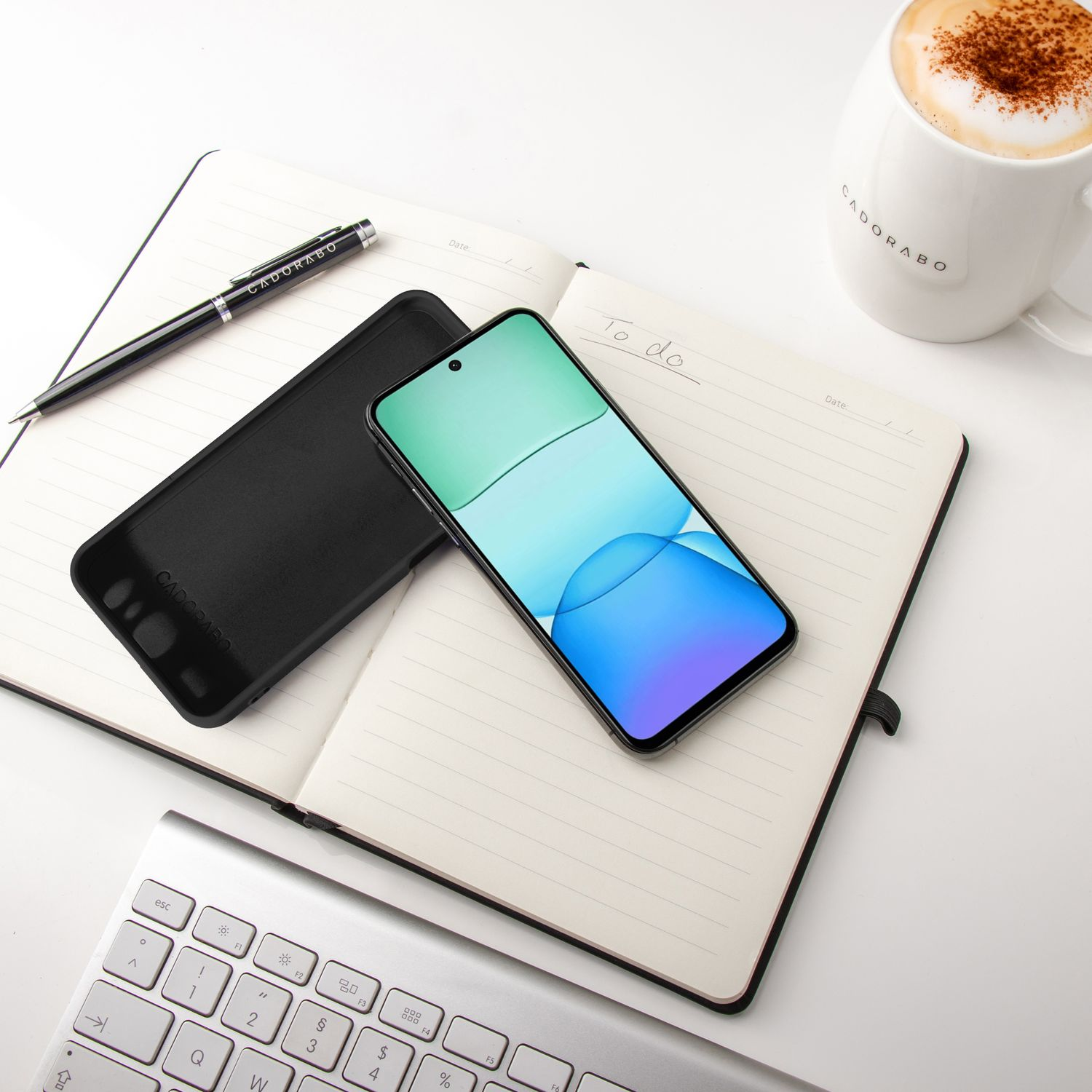 Notizbuch, Stift, Telefon und Kaffeetasse auf weißer Oberfläche. Das Telefon zeigt einen Farbverlauf.