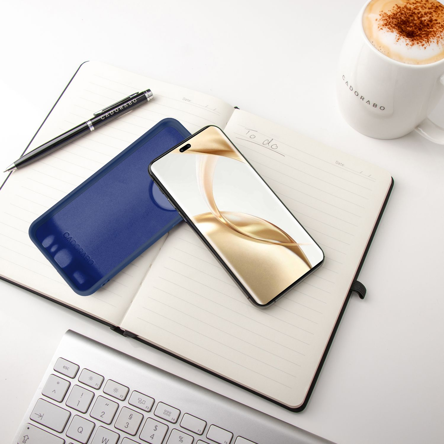 Notizbuch mit Telefon und Hülle. Stift und Becher liegen auf dem weißen Schreibtisch.