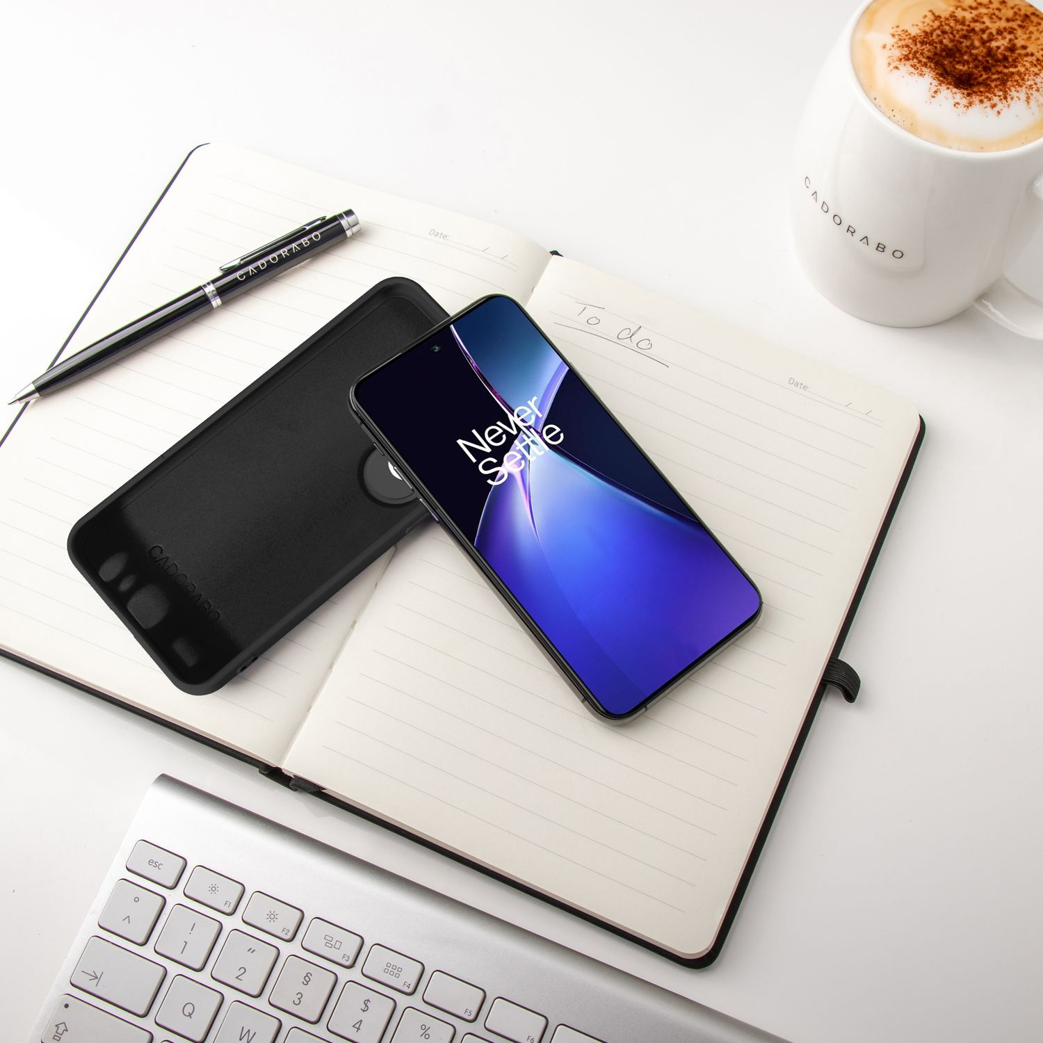 Ein Telefon mit blauem und lila Bildschirm, eine schwarze Hülle, Stift, Notizbuch und Kaffeetasse auf einem weißen Schreibtisch.