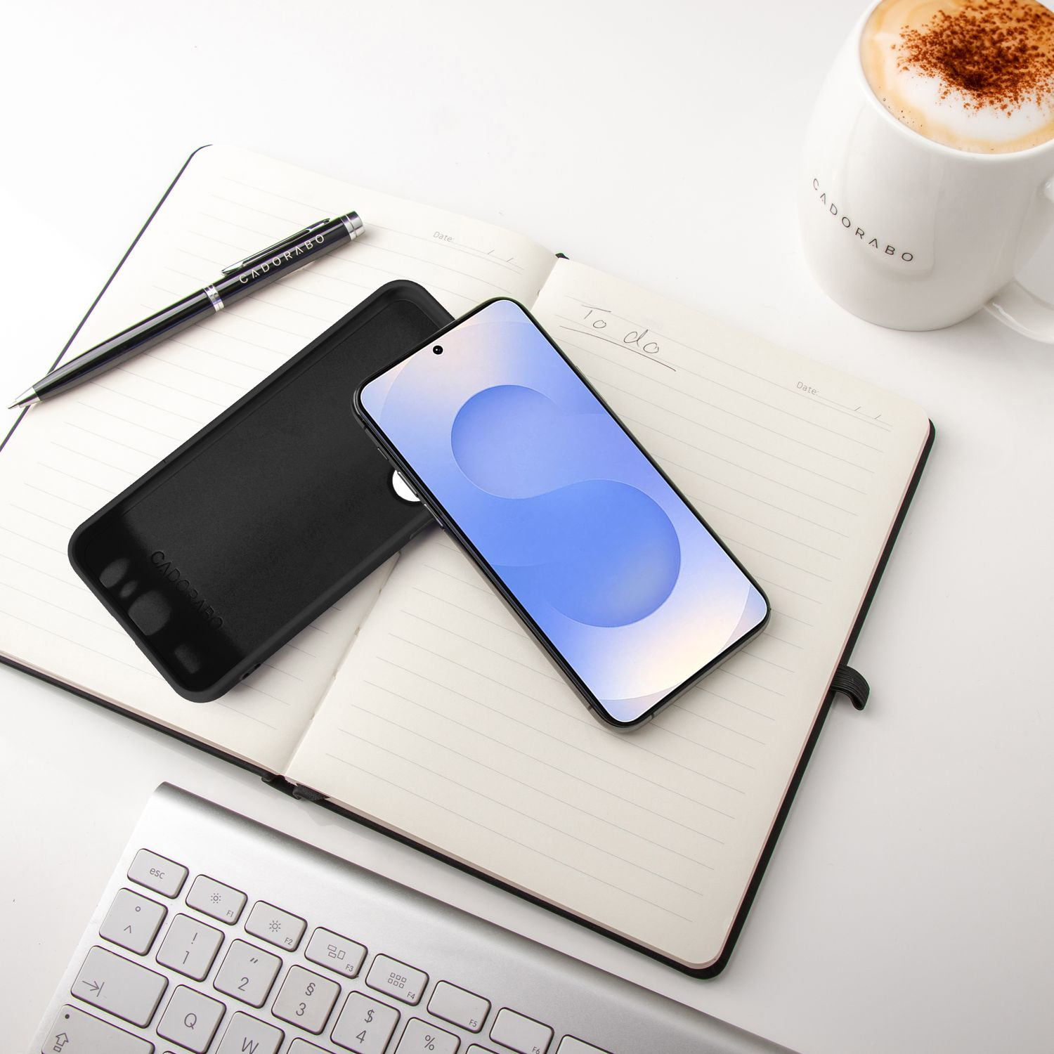 Smartphone, Hülle, Stift und Kaffee auf einem Notizbuch und Tastatur auf weißer Oberfläche.