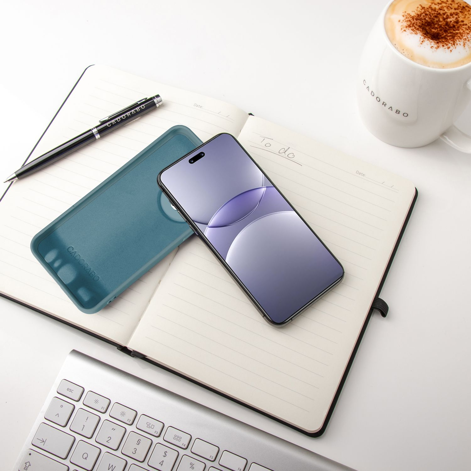Notizbuch mit Telefon und Hülle darauf. Auf dem Tisch liegen ein Stift und eine Tasse Kaffee.