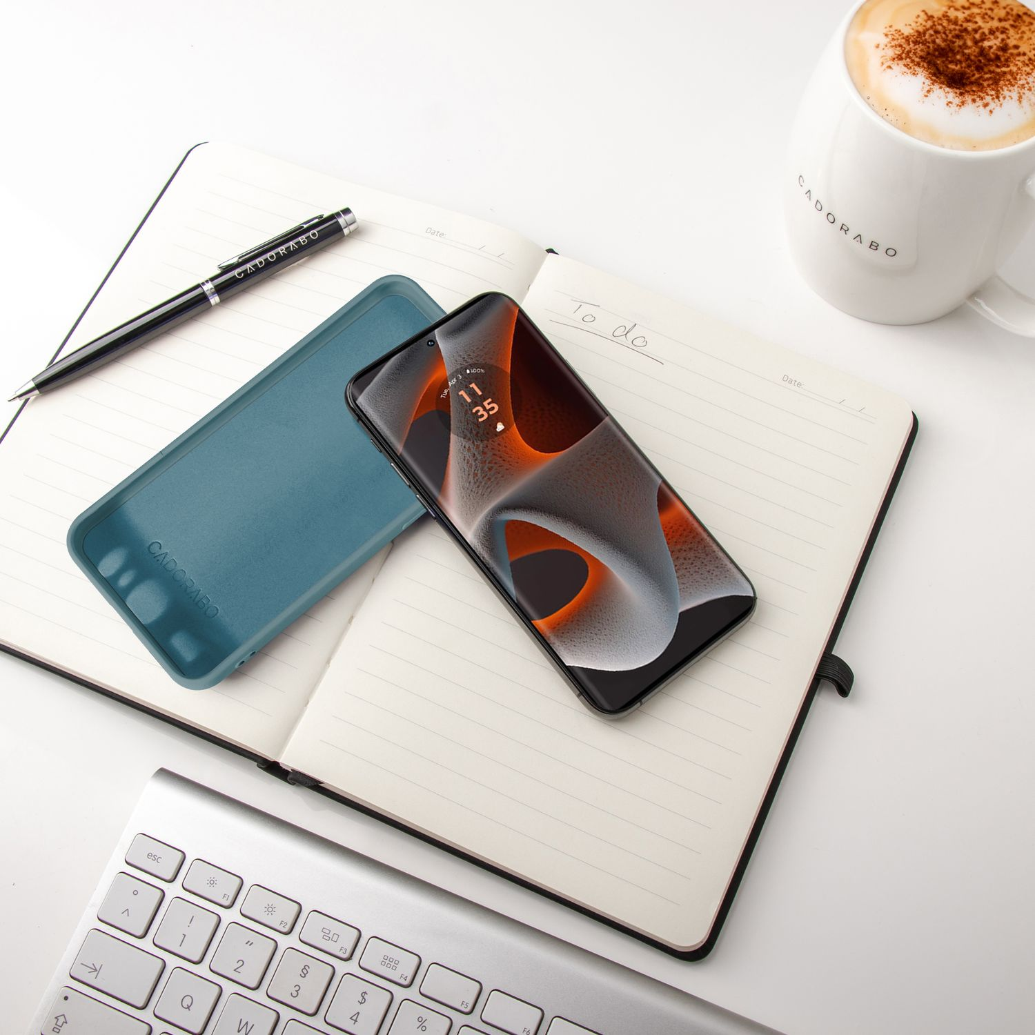 Notizbuch, Stift, Telefon und Hülle auf weißem Schreibtisch. Telefon zeigt Zeit und Datum. Cappuccino und Tastatur.