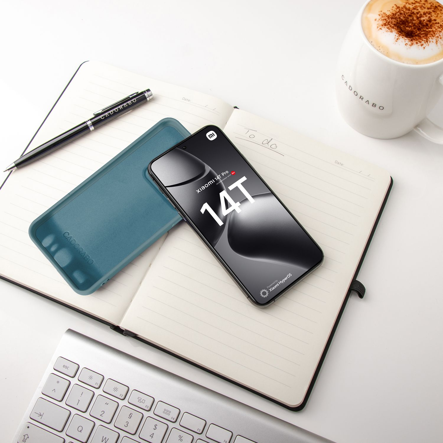 Ein Telefon, eine Hülle, ein Stift und eine Kaffeetasse liegen auf einem Notizbuch. Eine Tastatur ist im Hintergrund.