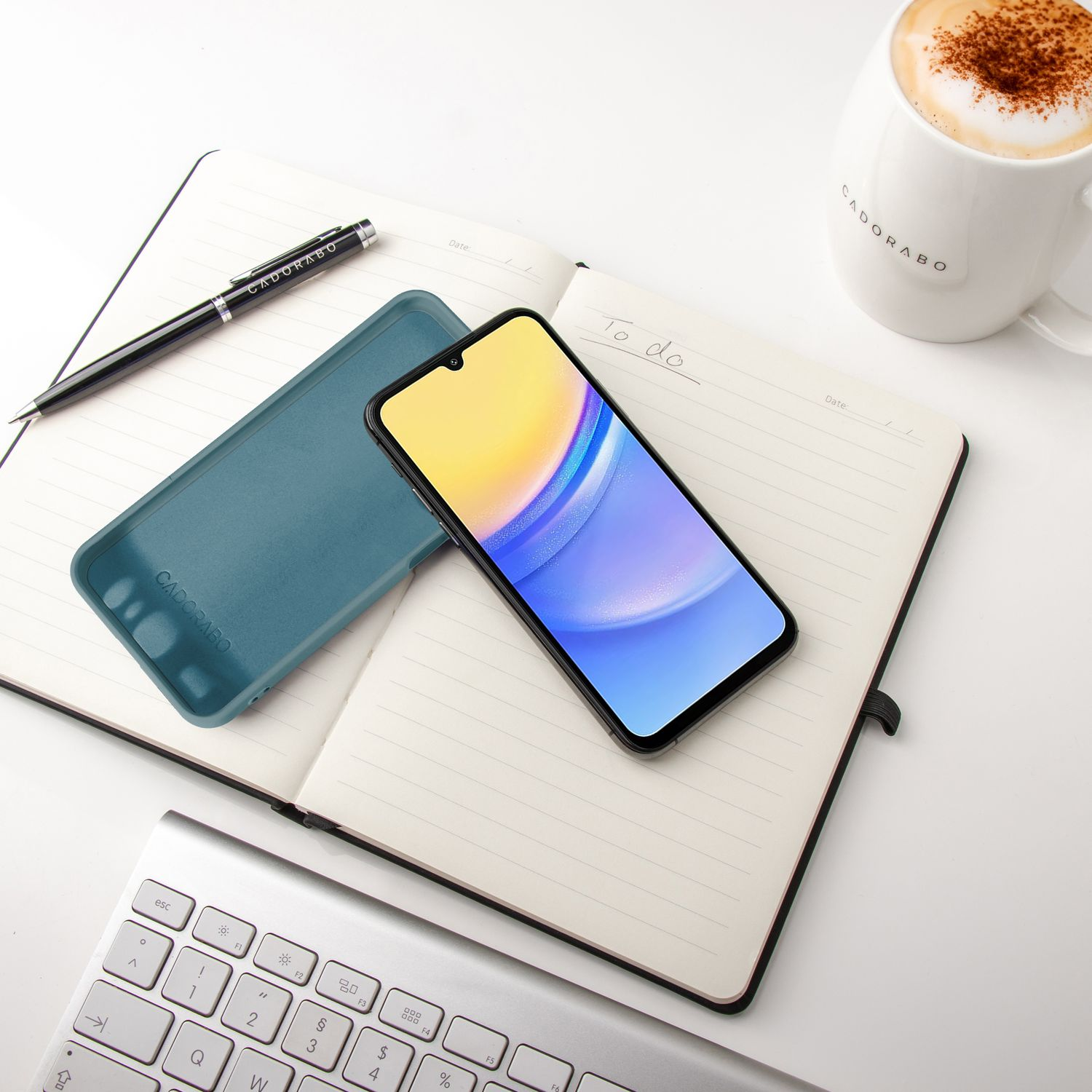 Ein Telefon mit blau-gelbem Bildschirm, ein Notizbuch, Stift und eine Kaffeetasse auf weißer Oberfläche.