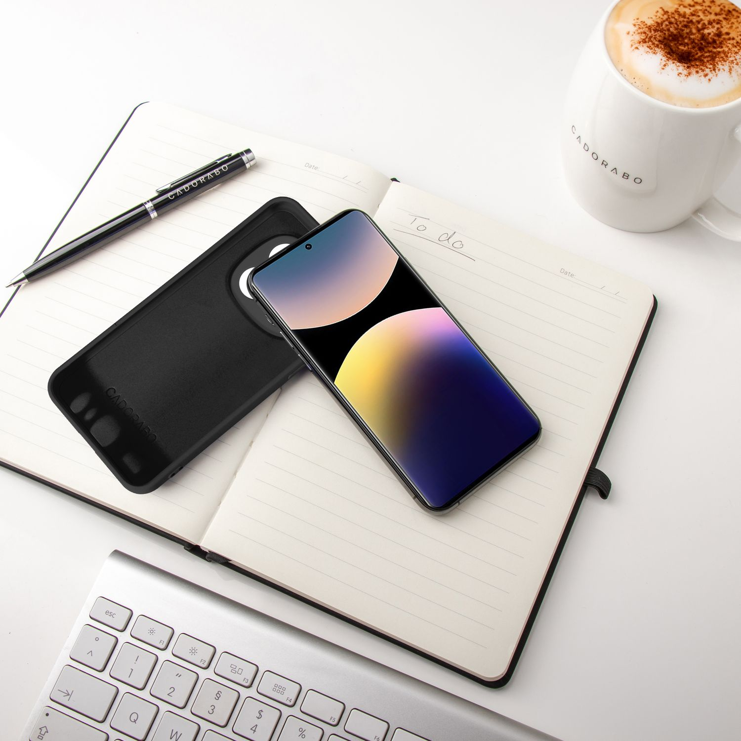 Ein Telefon in einer Hülle liegt auf einem Notizbuch mit Stift und Kaffeetasse auf einem weißen Schreibtisch.