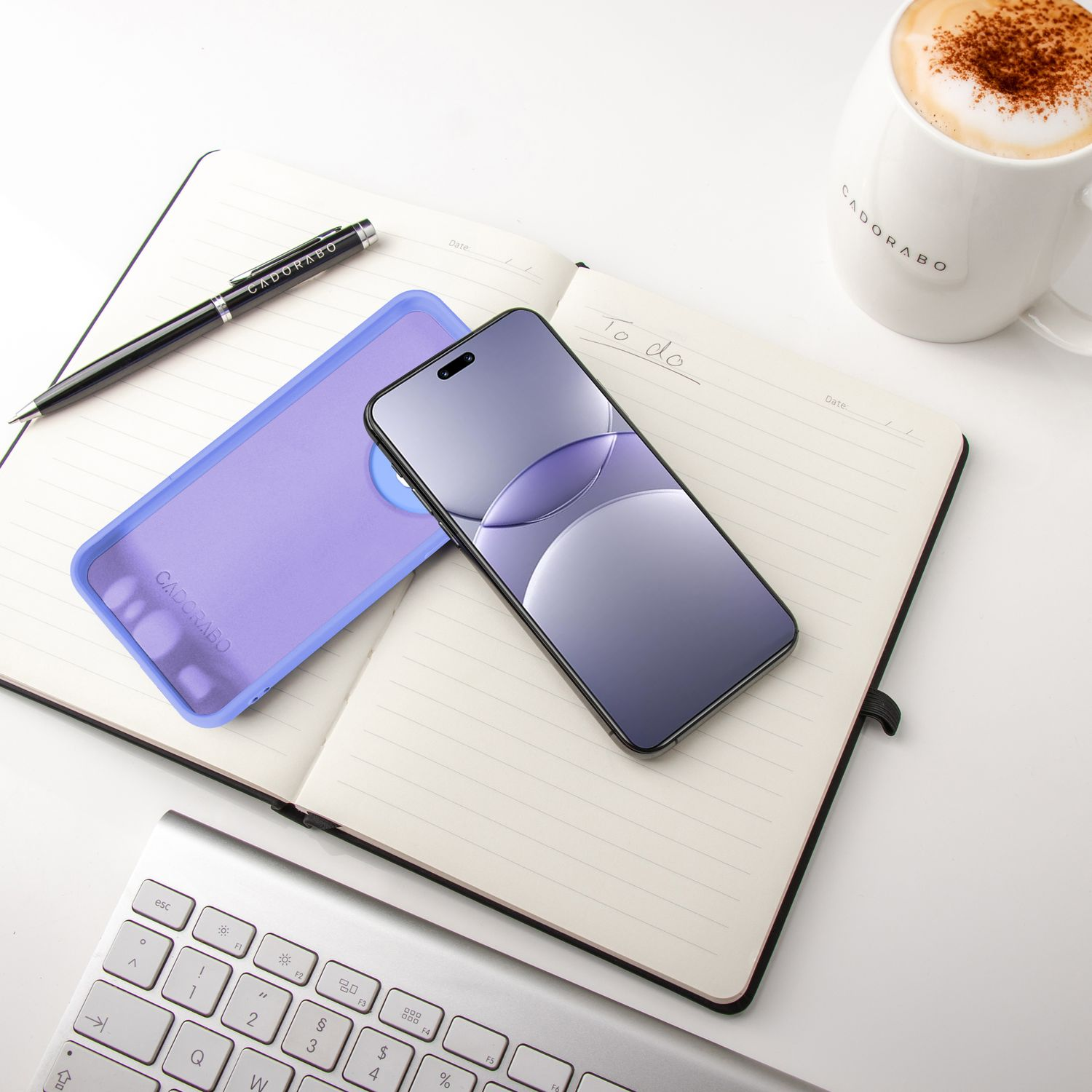 Notizbuch, Stift und Handyhülle mit einem Telefon oben drauf. Kaffeetasse im Hintergrund, zusammen mit einer Tastatur.