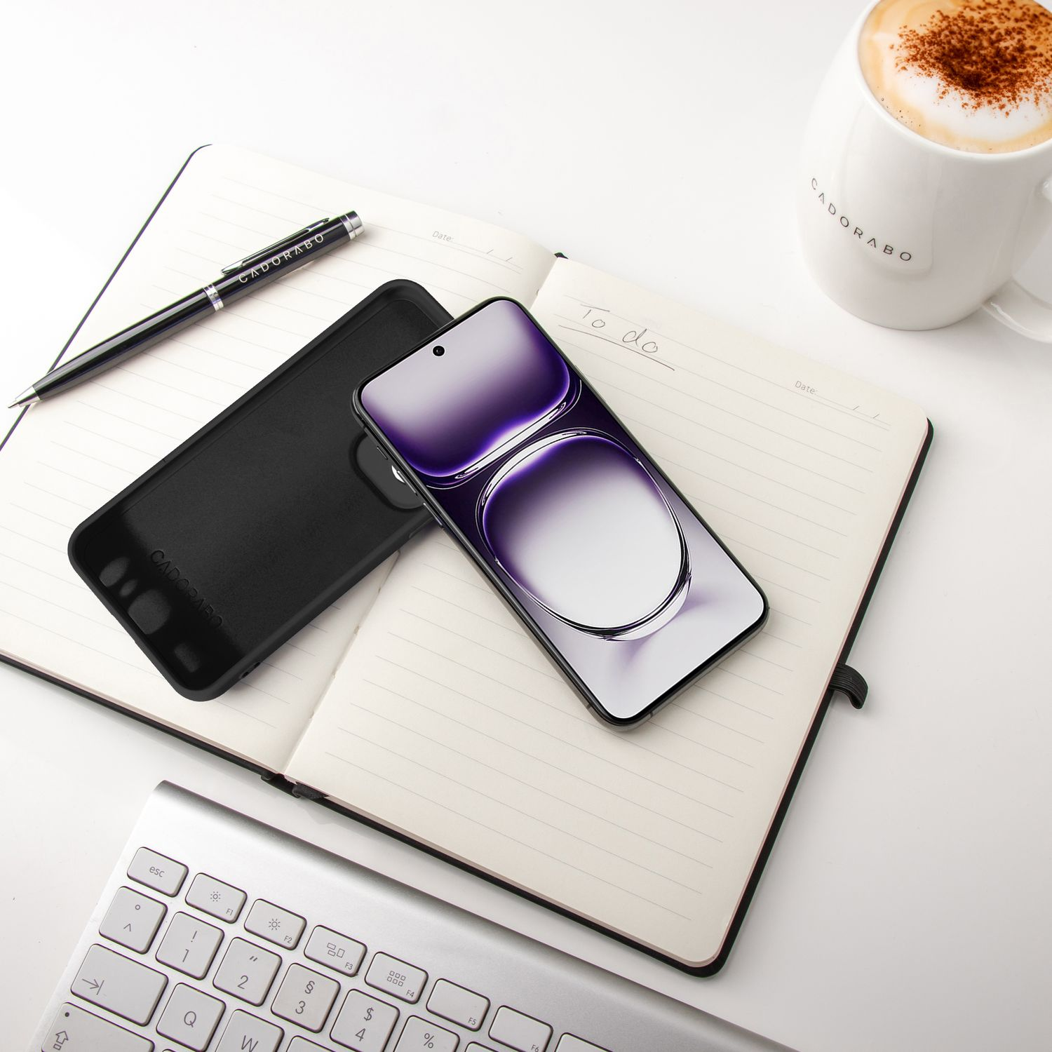 Schwarzes Telefon auf Notizbuch. Stift, Kaffee und Tastatur auf weißem Schreibtisch. Schwarze Handyhülle links.