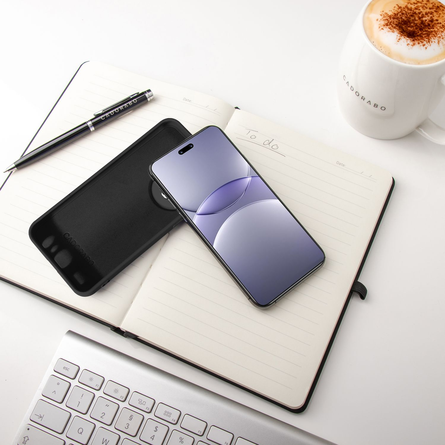 Telefon mit Hülle und Stift auf einem Notizbuch. Eine Tasse Latte und Tastatur sind ebenfalls im Bild.